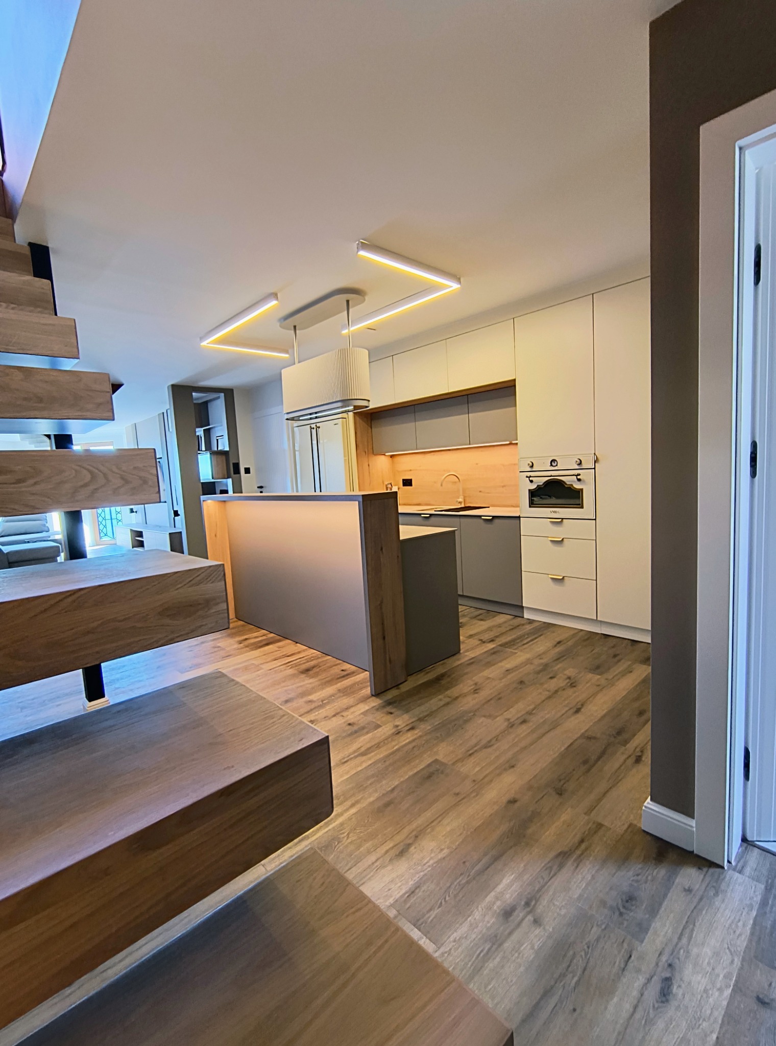 Nowoczesna kuchnia z wyspą i drewnianymi akcentami, widok z dołu schodów. Minimalistyczne oświetlenie LED i drewnopodobna podłoga tworzą spójną całość.