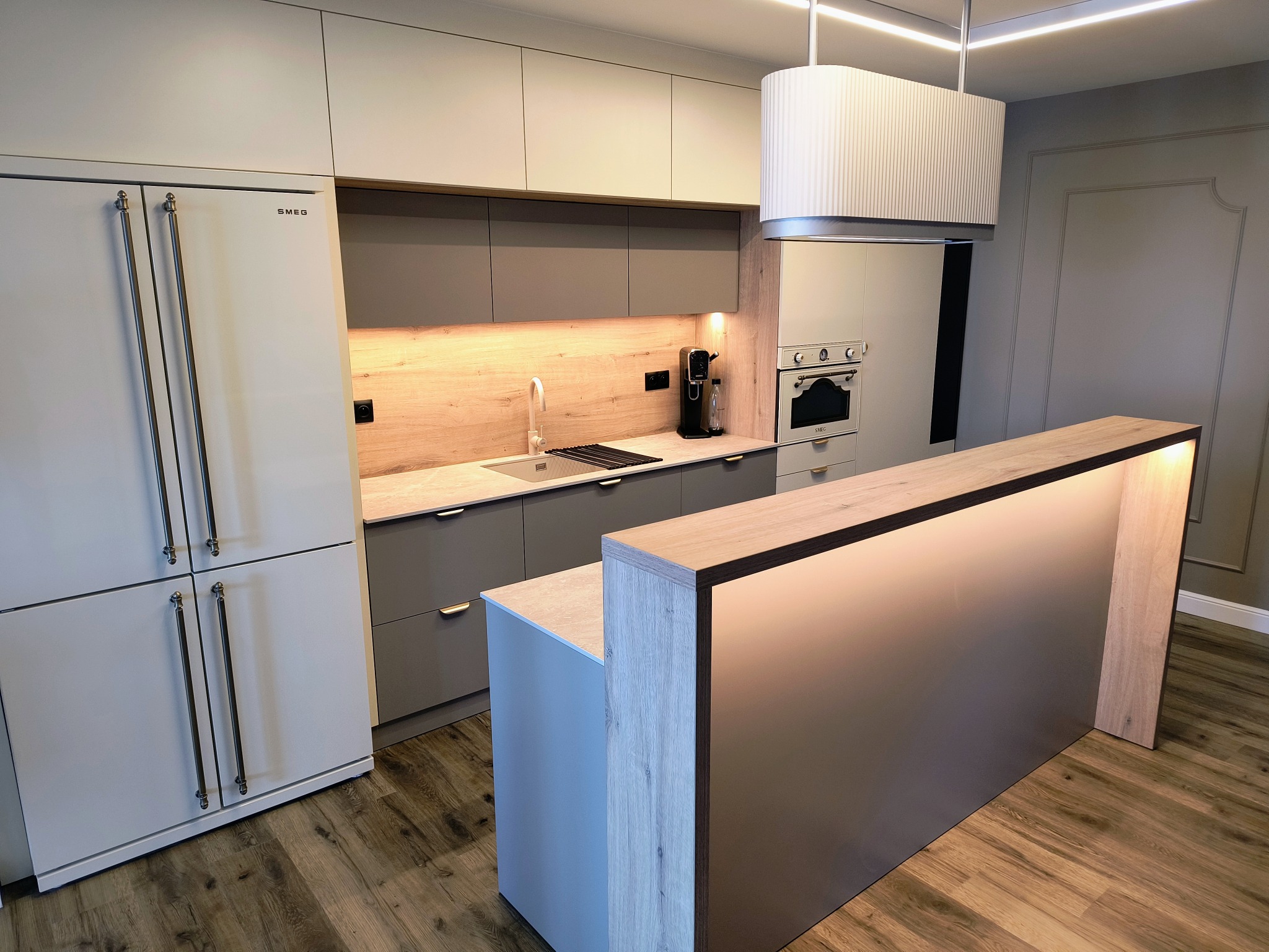 Nowoczesna kuchnia na wymiar w Ełku. Szare fronty szafek, drewniany blat i oświetlenie LED. Widoczna wyspa kuchenna i sprzęt AGD marki SMEG.
