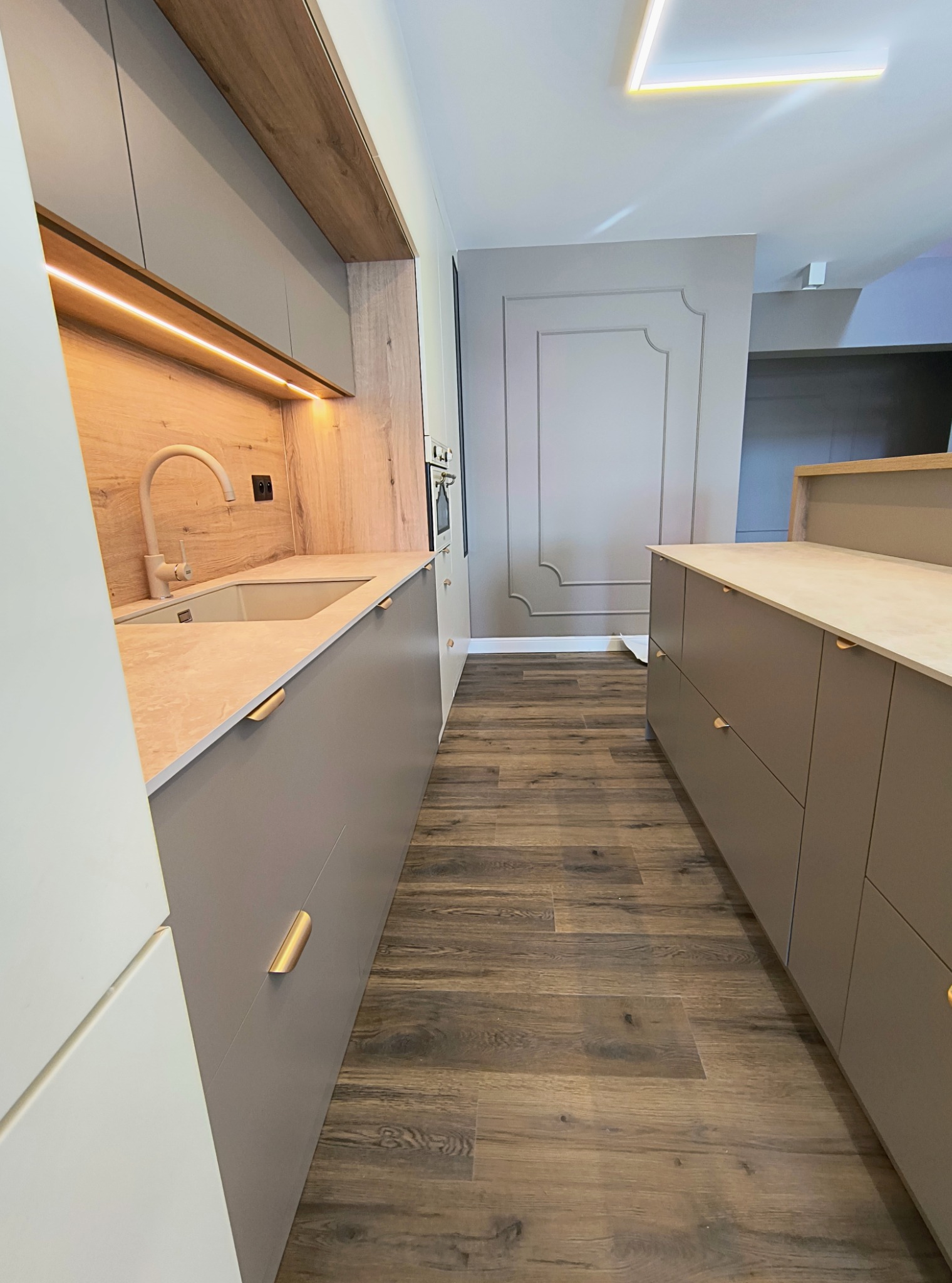 Nowoczesna kuchnia z szarymi szafkami, drewnianymi akcentami i oświetleniem LED. Uwagę przyciągają złote uchwyty i drewnopodobne panele podłogowe.