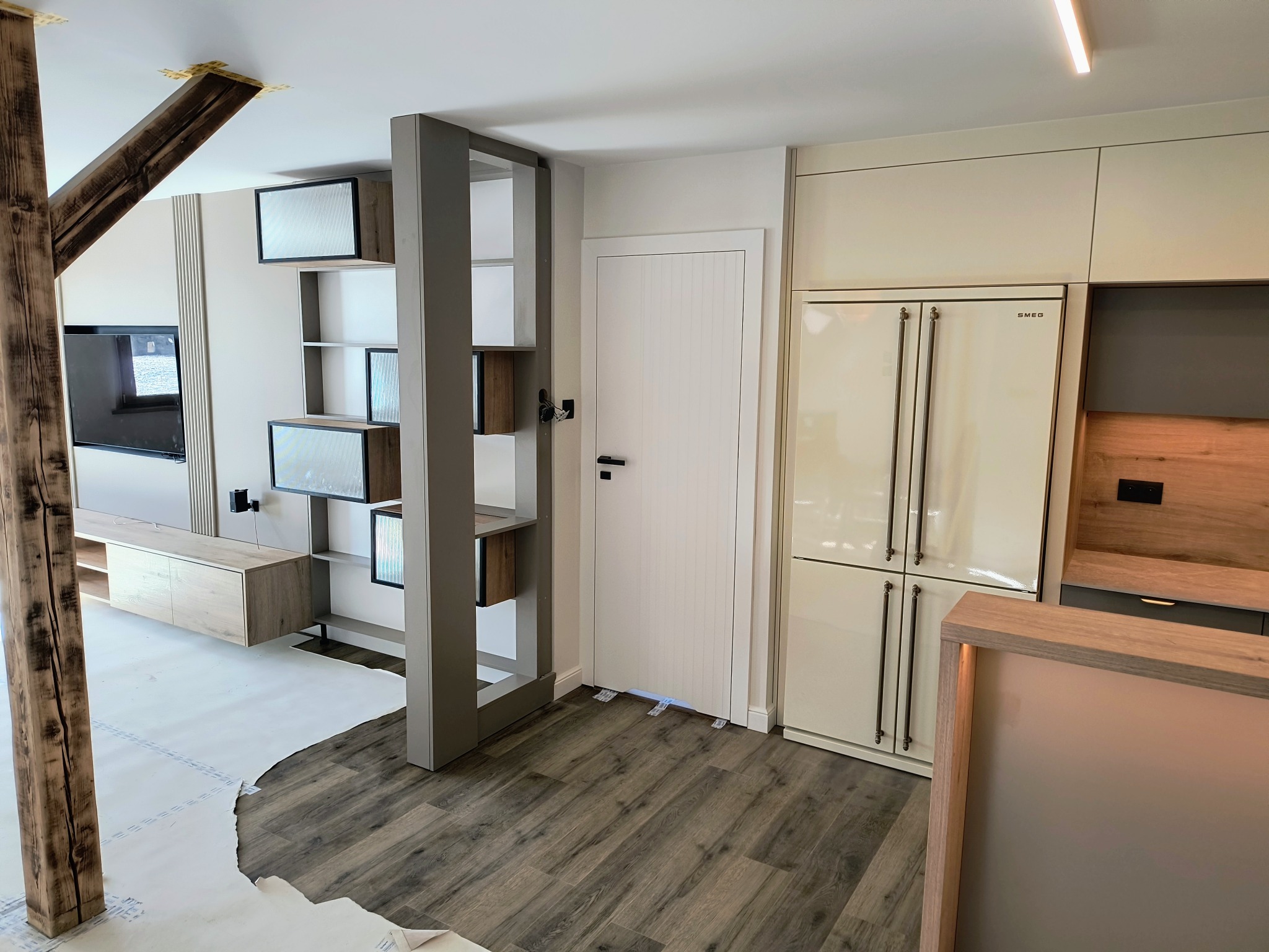 Nowoczesne wnętrze: kremowa zabudowa kuchenna SMEG, drewniany regał działowy, parkiet w odcieniach szarości. W tle drewniana belka i zabudowa RTV.