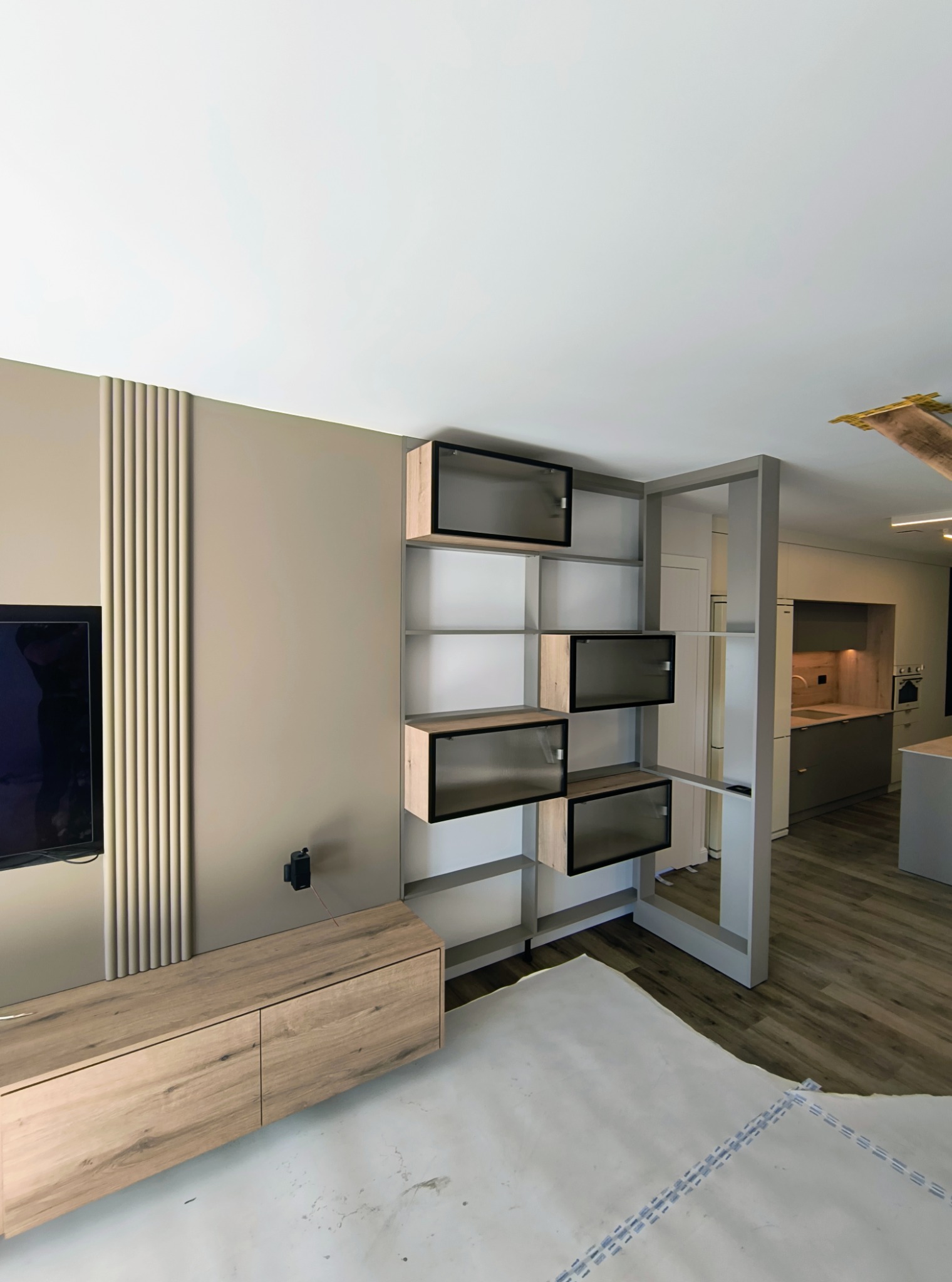 Nowoczesny salon z drewnianą szafką RTV, regałem z przeszklonymi drzwiczkami i widokiem na kuchnię z szarymi frontami. Podłoga wyłożona panelami.