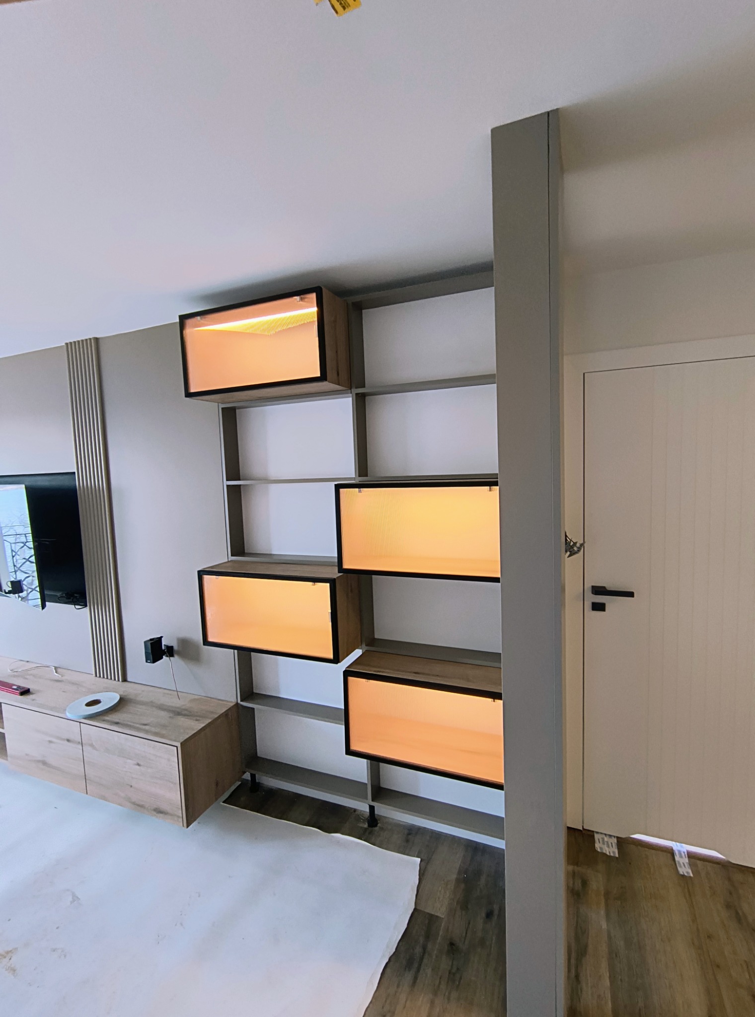 Nowoczesna szafa wnękowa z podświetlanymi półkami i elementami drewnopodobnymi, zamontowana w jasnym wnętrzu. Widoczny fragment mebla RTV i drzwi.