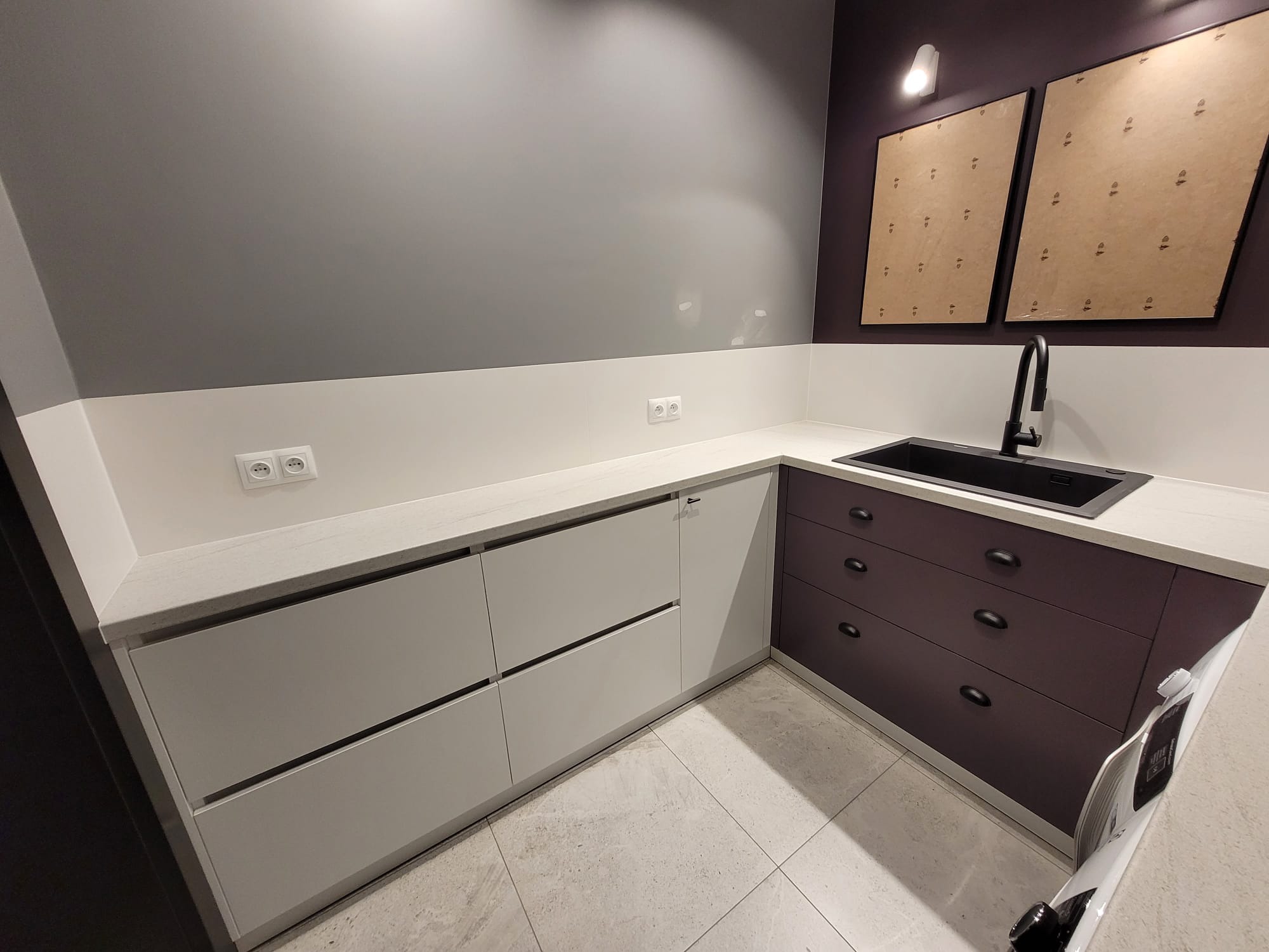 Nowoczesna kuchnia z białymi i fioletowymi szafkami, czarnym zlewem i minimalistyczną armaturą. Dwa obrazy w ramkach na ścianie dodają charakteru.