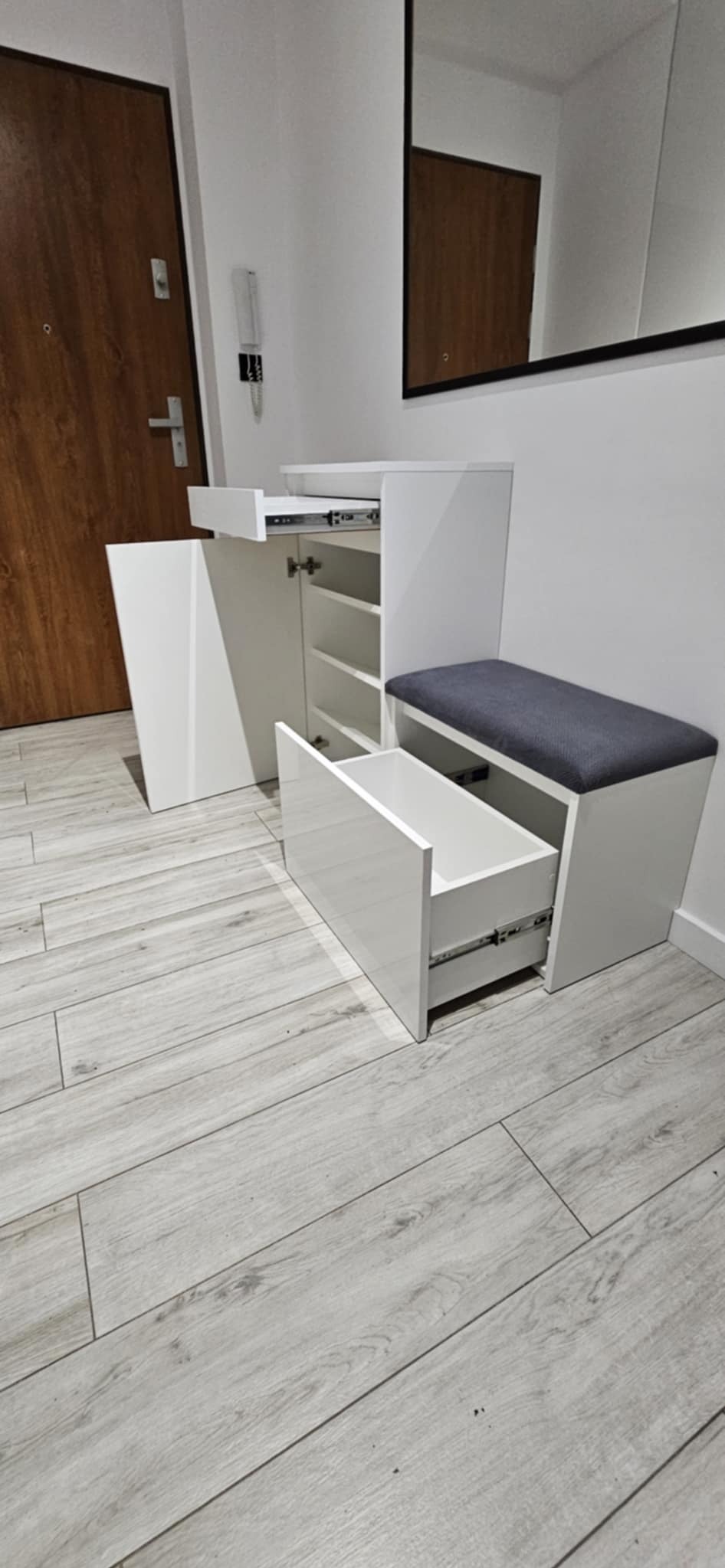 Biała szafka na buty z siedziskiem i wysuwanymi szufladami, widoczna w przedpokoju na tle drewnianych drzwi i lustra. Szuflady częściowo otwarte, ukazujące mechanizm.