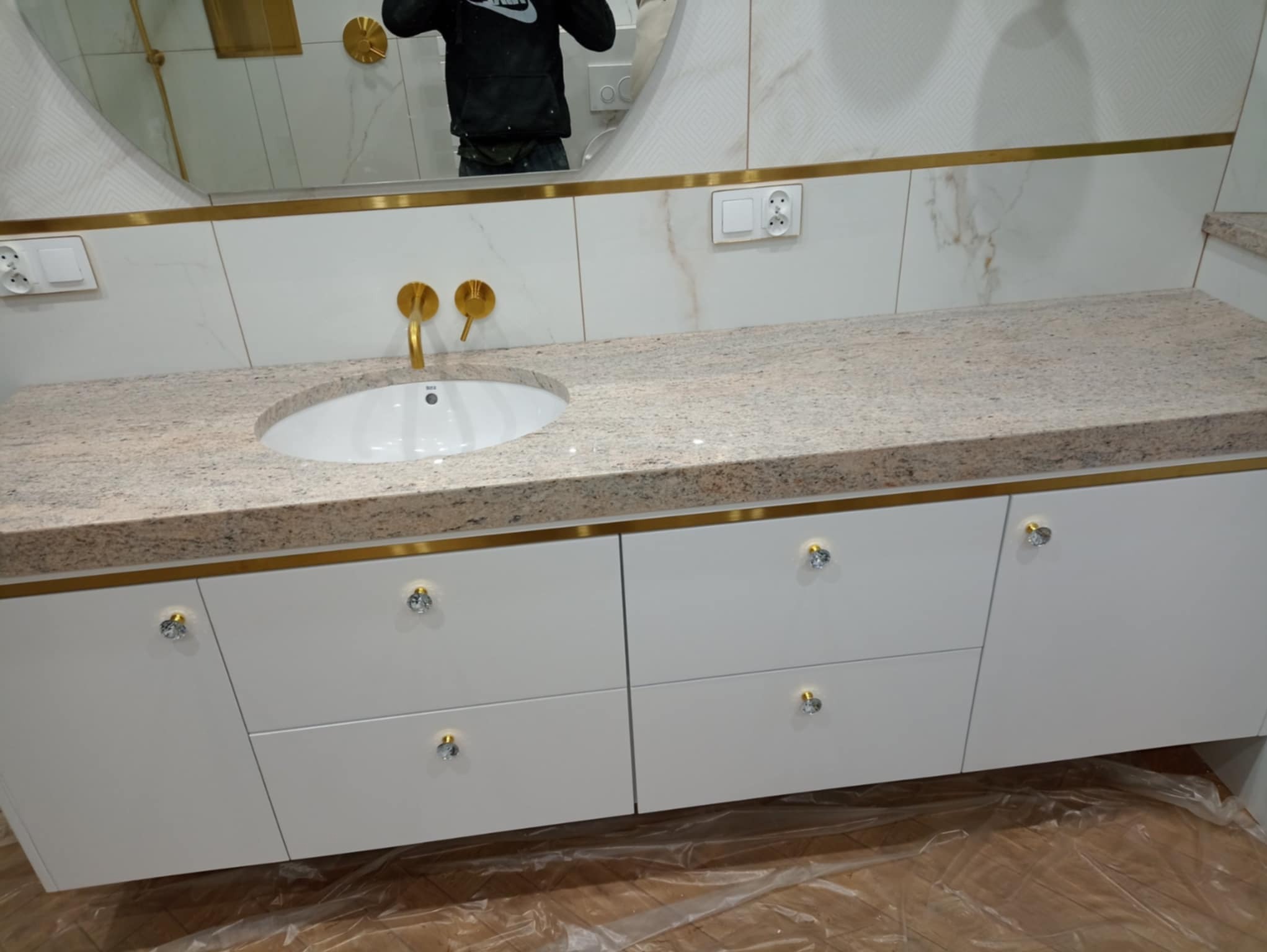 Biała szafka łazienkowa z blatem z jasnego kamienia, złotymi detalami i kryształowymi gałkami, odbicie osoby w lustrze. Nowoczesny design.