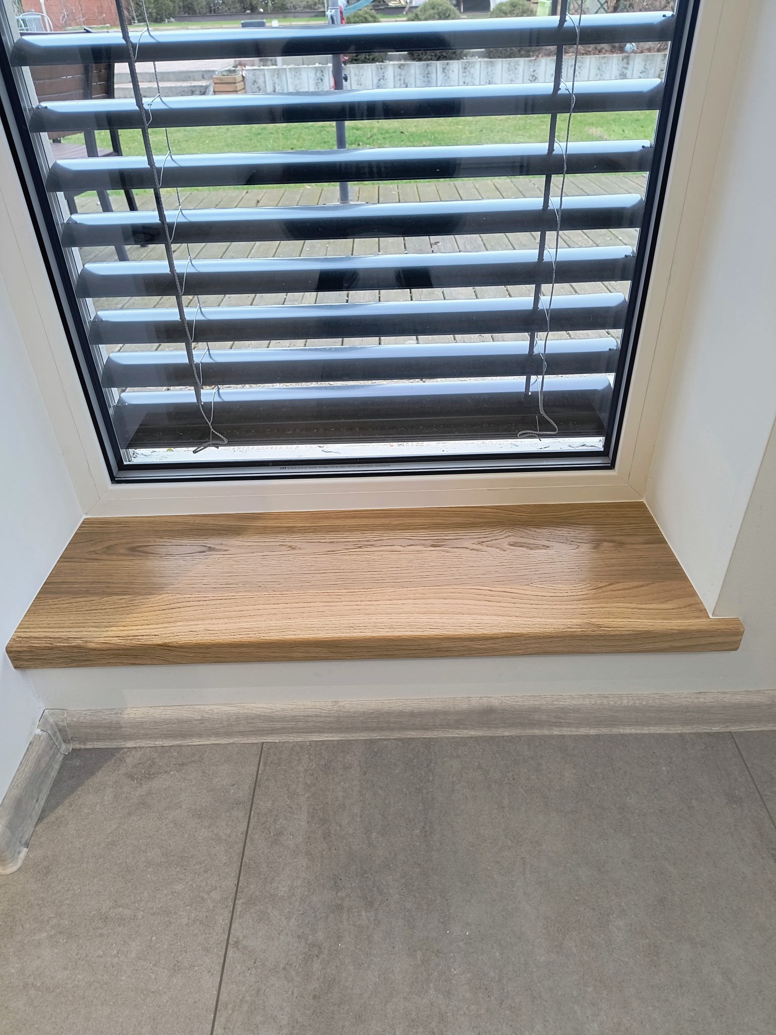 Jasny, dębowy parapet wewnętrzny z zaokrąglonym brzegiem, zamontowany pod oknem z żaluzjami poziomymi. Szara podłoga z płytek i białe ściany.