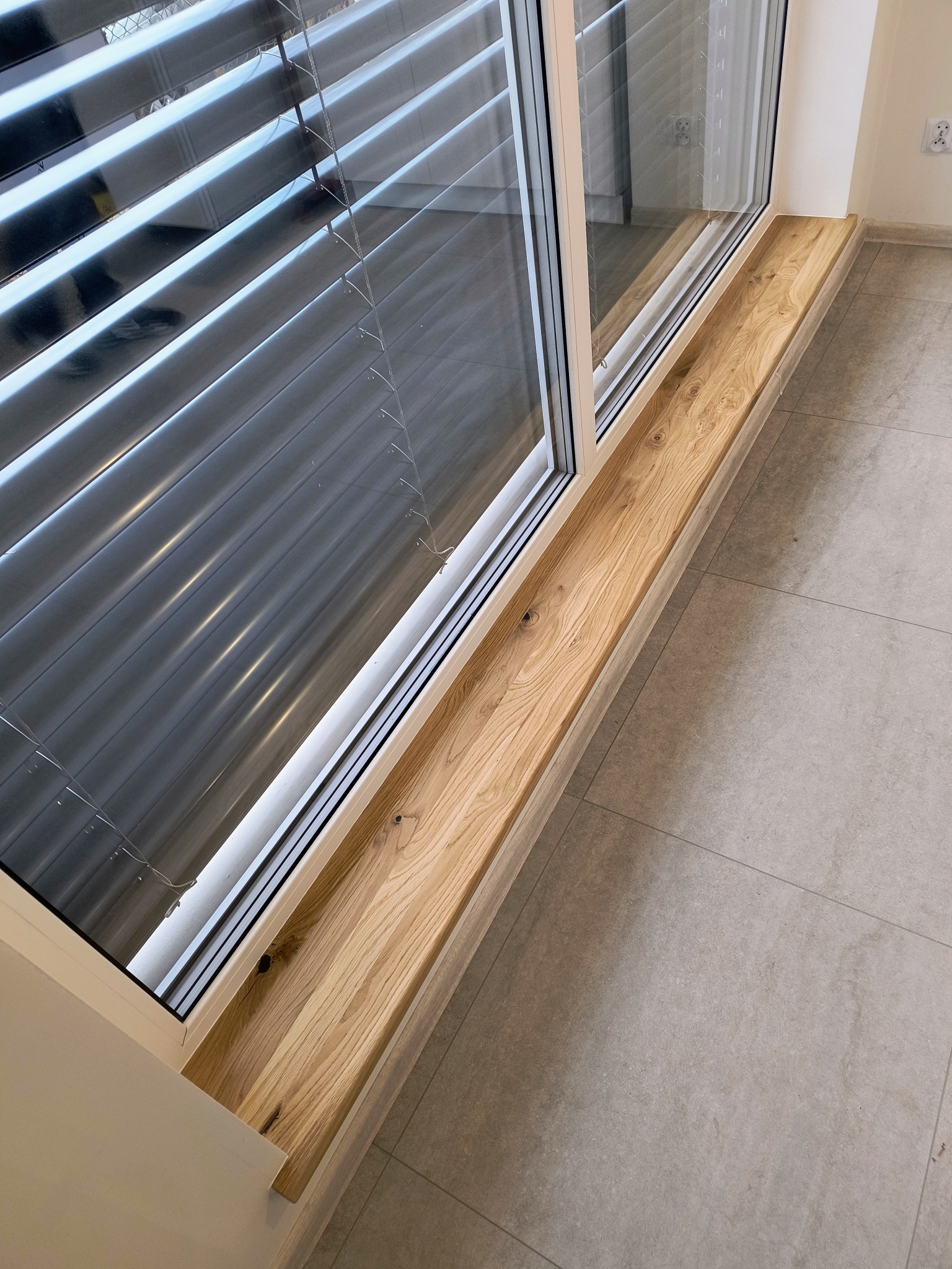 Jasny, drewniany parapet z wyraźnym usłojeniem, wkomponowany w nowoczesne okno z żaluzjami, na tle szarej, kwadratowej podłogi z płytek. Detal wnętrza.