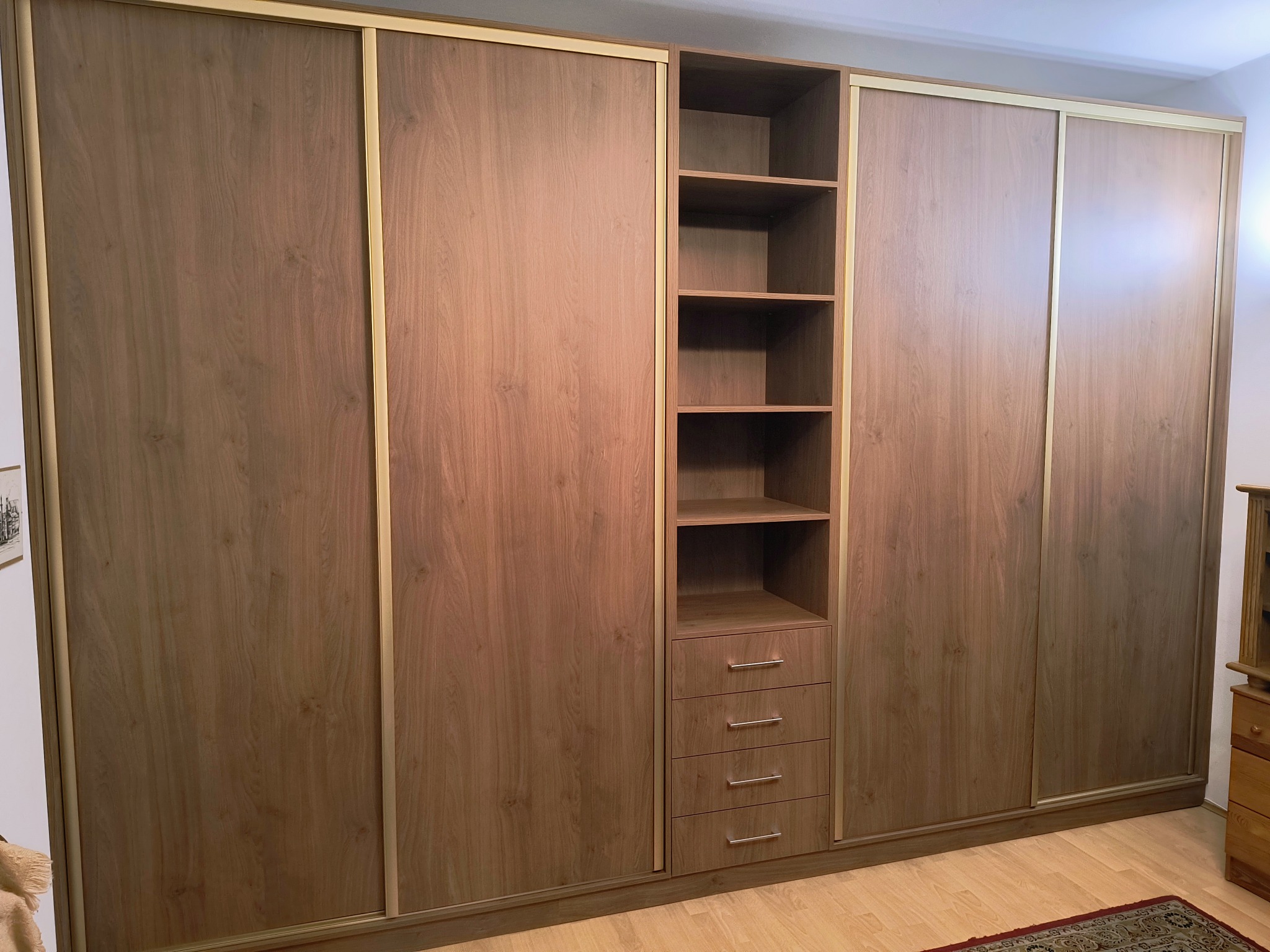 Drewniana szafa wnękowa z przesuwnymi drzwiami i złotymi detalami. Widoczne półki i szuflady. Ciepłe oświetlenie podkreśla naturalny rysunek drewna.