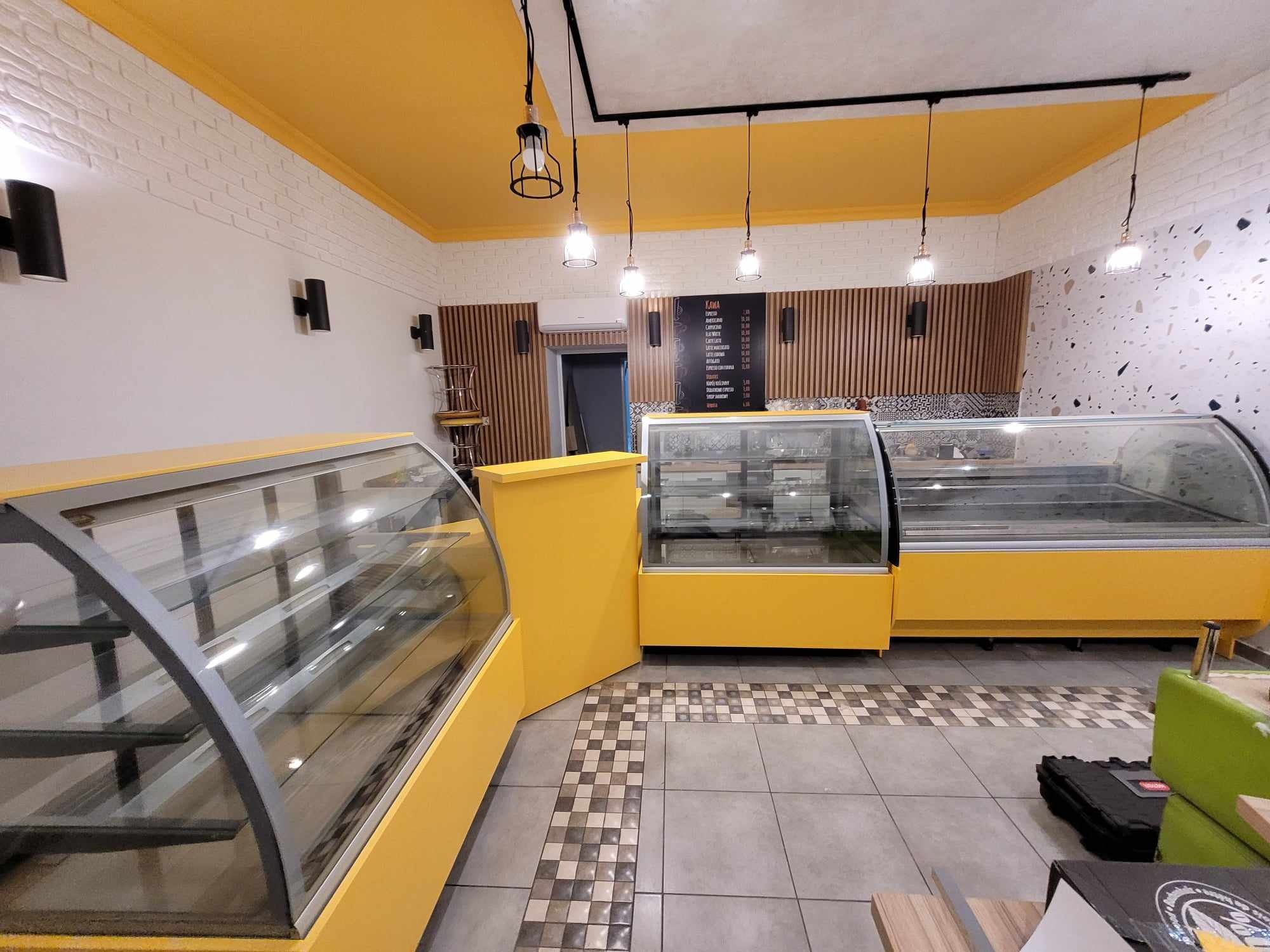 Żółte lady chłodnicze w nowoczesnym lokalu z ceglaną ścianą i drewnianymi panelami. Nad nimi wisi menu i lampy. Podłoga wyłożona jest szarymi i wzorzystymi płytkami.