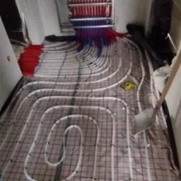 Instalacja ogrzewania podłogowego z rozdzielaczem w pomieszczeniu z poddaszem, rurki białe, mocowane do siatki, widoczny rozdzielacz z rurkami czerwonymi i niebieskimi.