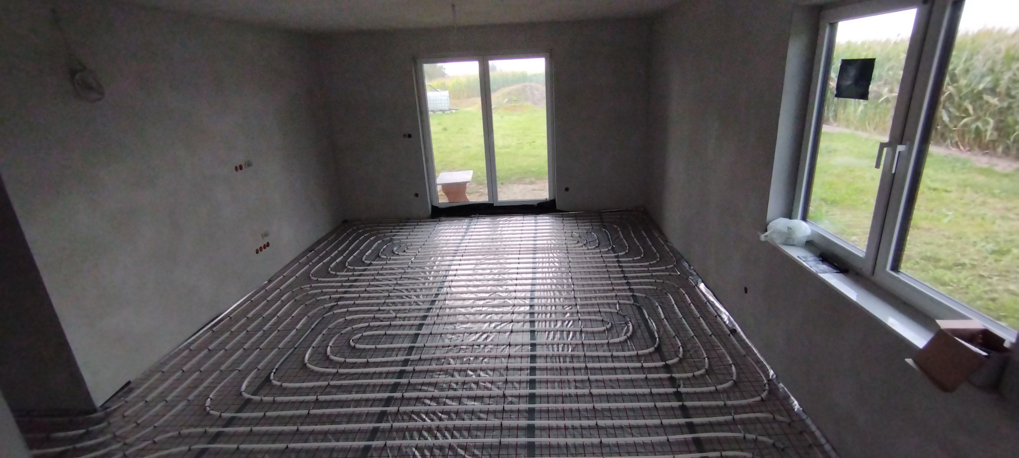 Szare rury ogrzewania podłogowego ułożone w pokoju z oknem i surowymi ścianami. Etap instalacji przed wylewką. Widok na zewnątrz przez okno.