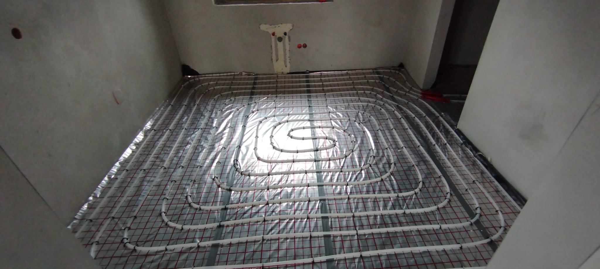 Białe rury ogrzewania podłogowego ułożone na folii w surowym wnętrzu. Instalacja w kształcie spirali, widoczne uchwyty mocujące. Przygotowanie do wylewki.