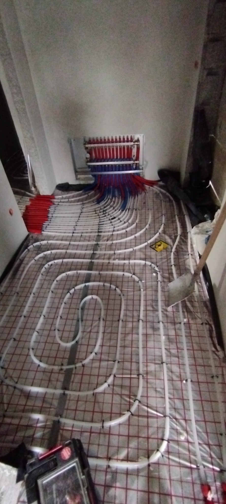 Instalacja ogrzewania podłogowego z rozdzielaczem w pomieszczeniu z poddaszem, rurki białe, mocowane do siatki, widoczny rozdzielacz z rurkami czerwonymi i niebieskimi.