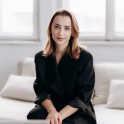 Portret kobiety w czarnym garniturze, siedzącej na jasnej kanapie z laptopem w tle, w jasnym wnętrzu z oknem. Uśmiechnięta, z delikatnym makijażem i czerwoną stylizacją paznokci.