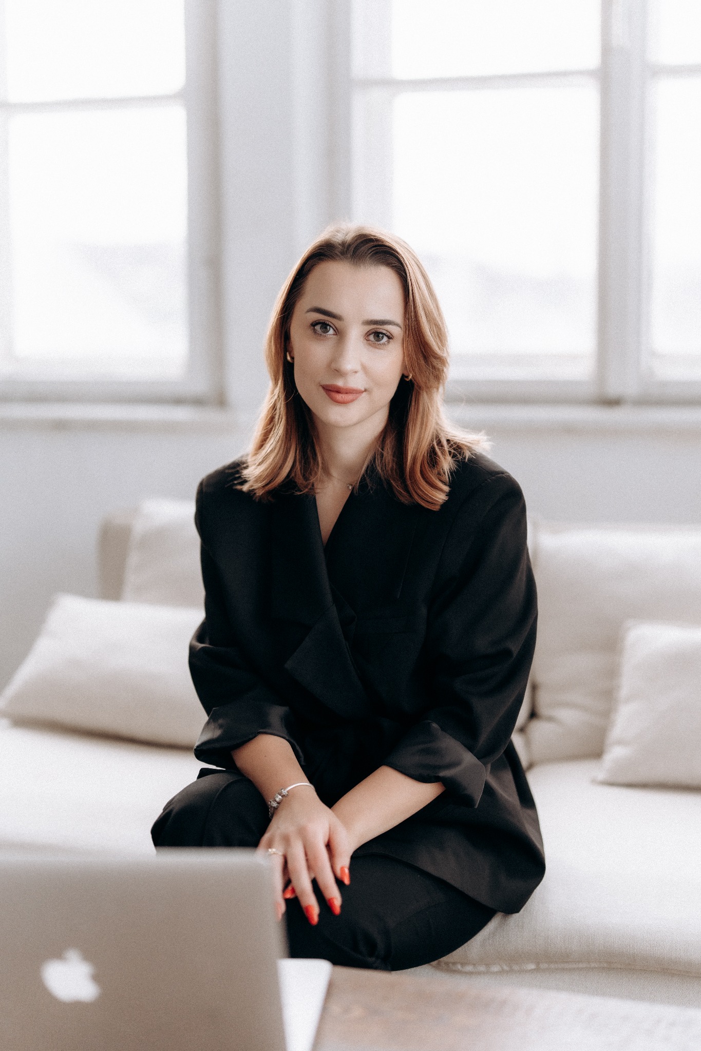 Portret kobiety w czarnym garniturze, siedzącej na jasnej kanapie z laptopem w tle, w jasnym wnętrzu z oknem. Uśmiechnięta, z delikatnym makijażem i czerwoną stylizacją paznokci.