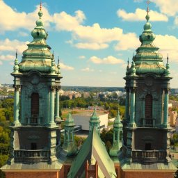 Kamil Cieslak - Panorama miasta z perspektywy drona, z bliska ukazująca dwie wieże kościoła o zielonych kopułach na tle błękitnego nieba z obłokami.