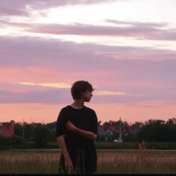 Kamil Cieslak - Młody mężczyzna w czerni, stojący na tle różowo-fioletowego nieba o zmierzchu, patrzy w bok. W tle widoczne zabudowania i trawa.