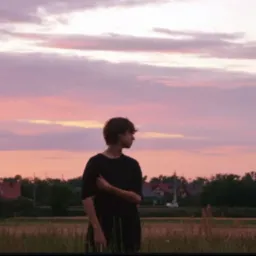 Młody mężczyzna w czerni, stojący na tle r&oacute;żowo-fioletowego nieba o zmierzchu, patrzy w bok. W tle widoczne zabudowania i trawa.