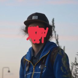 Kamil Cieslak - Młody mężczyzna w czapce i niebieskiej bluzie z aparatem na szyi, na tle nieba o zmierzchu. Widoczne trawy i lampa uliczna.