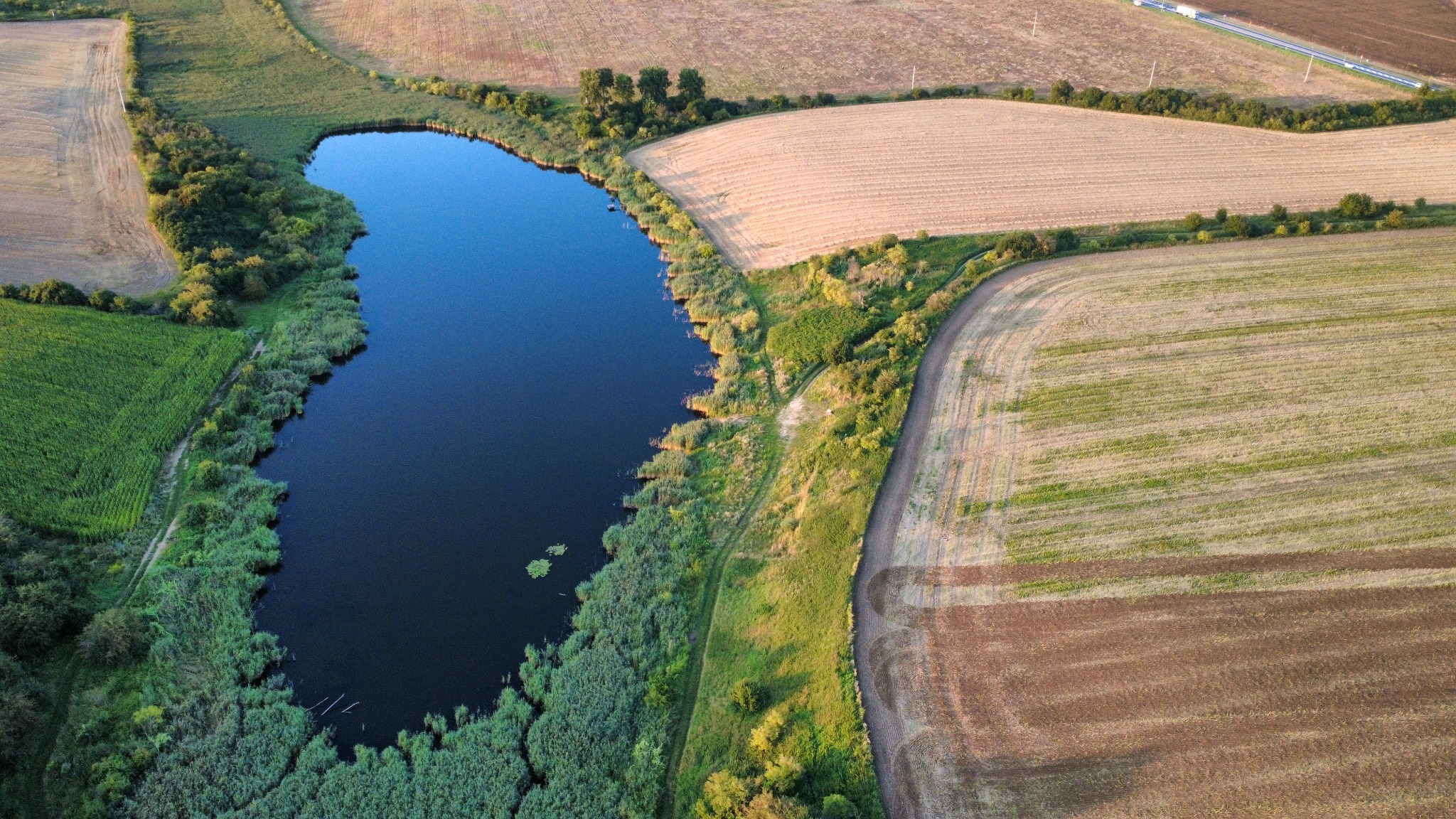 Panoramiczny widok z lotu ptaka na malownicze jezioro otoczone polami uprawnymi, z widoczną drogą w tle.