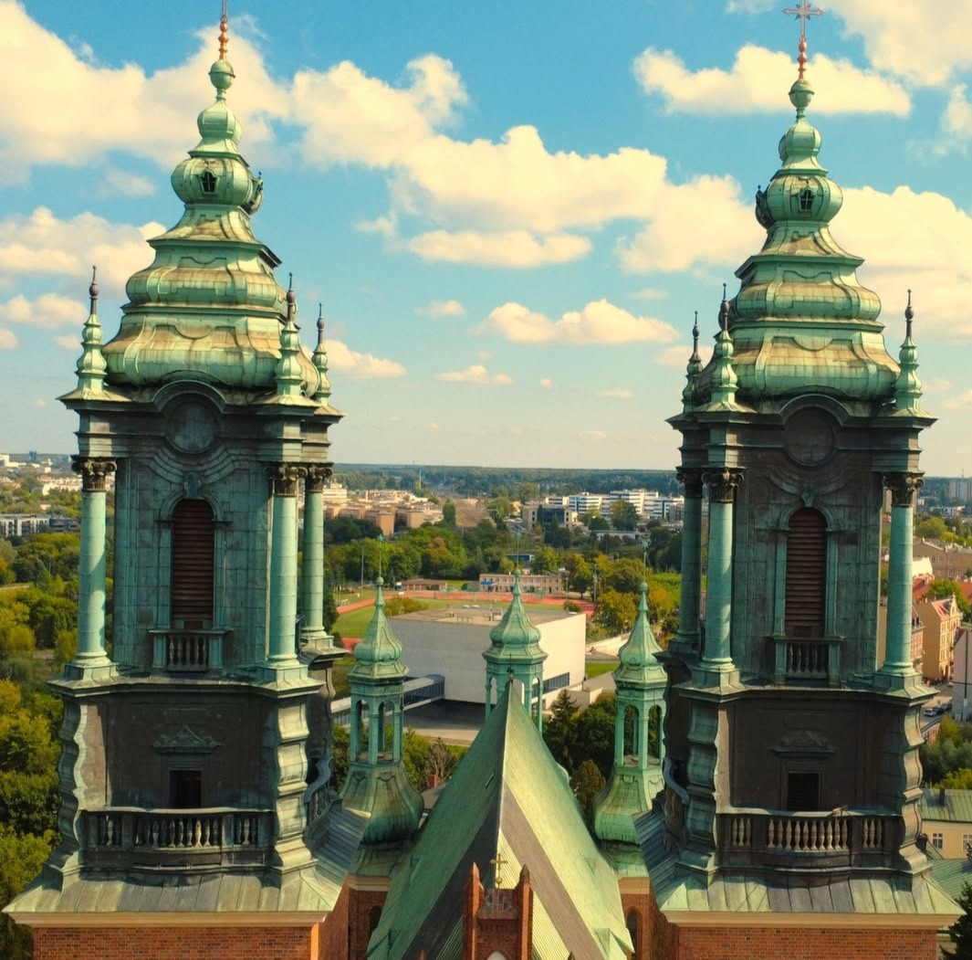 Panorama miasta z perspektywy drona, z bliska ukazująca dwie wieże kościoła o zielonych kopułach na tle błękitnego nieba z obłokami.