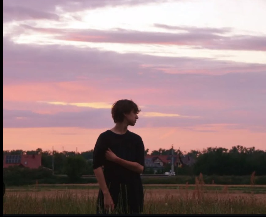 Młody mężczyzna w czerni, stojący na tle różowo-fioletowego nieba o zmierzchu, patrzy w bok. W tle widoczne zabudowania i trawa.