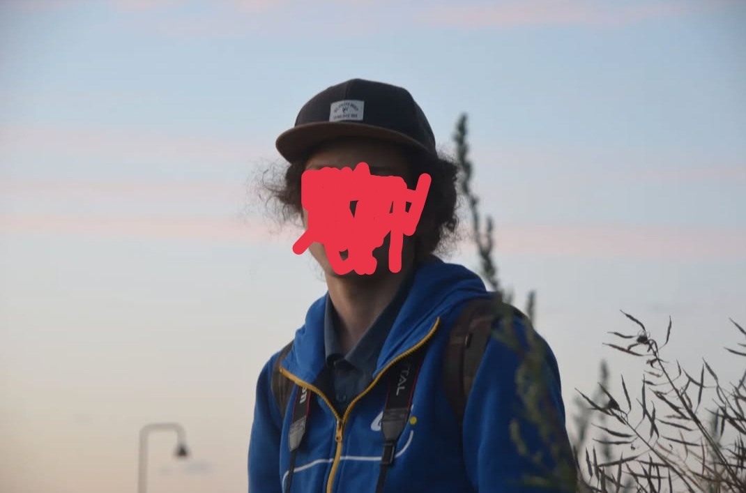 Młody mężczyzna w czapce i niebieskiej bluzie z aparatem na szyi, na tle nieba o zmierzchu. Widoczne trawy i lampa uliczna.