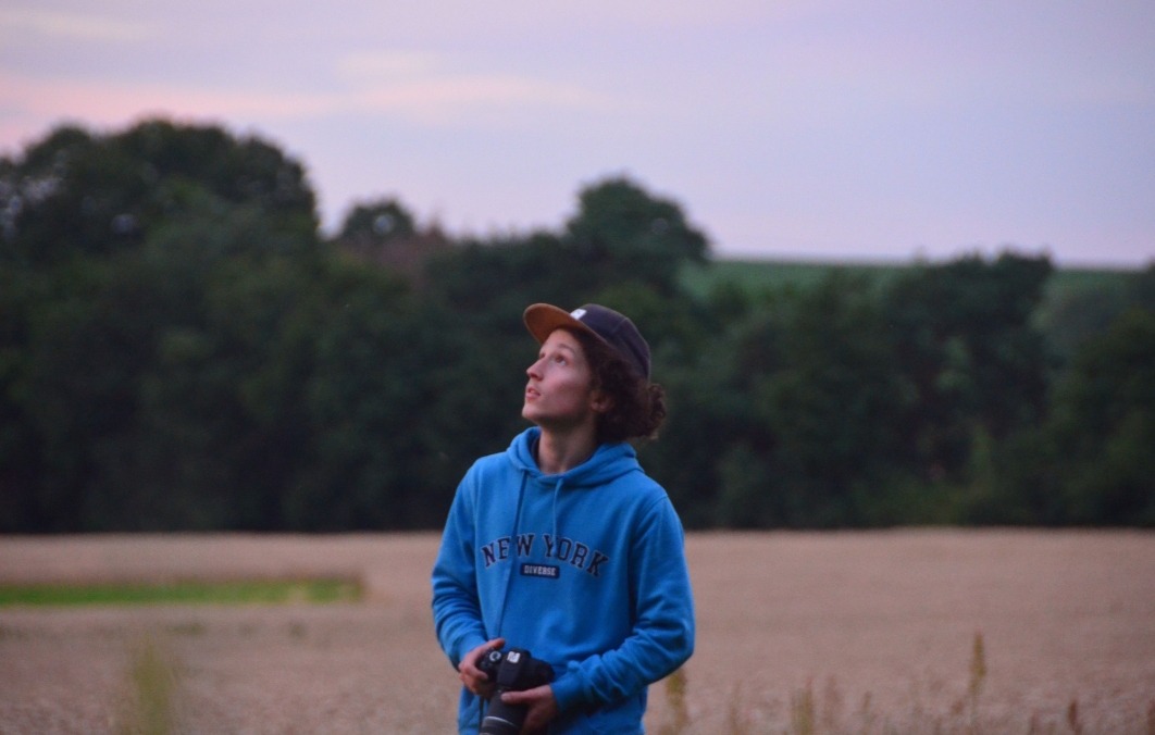 Młody fotograf z aparatem w dłoni, w bluzie z napisem 'New York', patrzy w górę na tle pola i lasu o zmierzchu. Ujęcie z dołu.