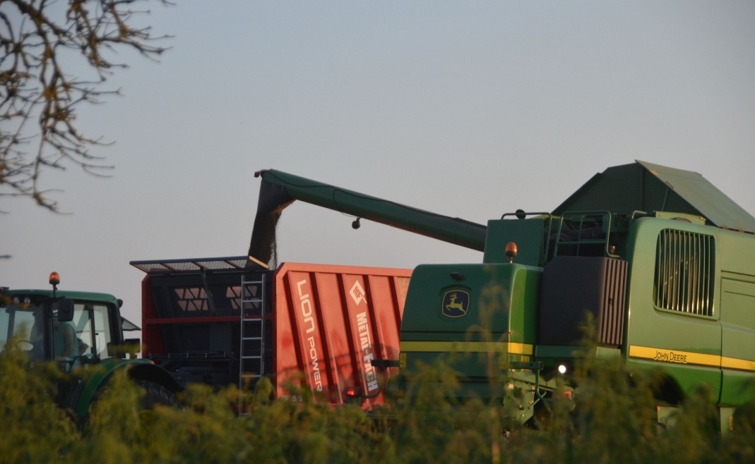 Zielony kombajn John Deere wysypuje ziarno do czerwonej przyczepy Lion Power w polu, widoczny także ciągnik. Scena żniw w słoneczny dzień.