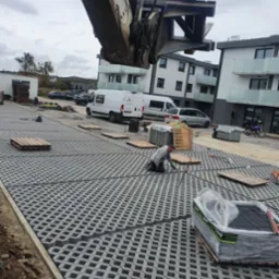 Montaż ażurowej nawierzchni parkingowej z użyciem ciężkiego sprzętu, widoczny pracownik w trakcie pracy, palety z kostką, budynki w tle.