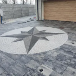 Szara kostka brukowa z dekoracyjnym wzorem kompasu w odcieniach szarości i bieli. W tle widoczny betonowy płot i brama garażowa.