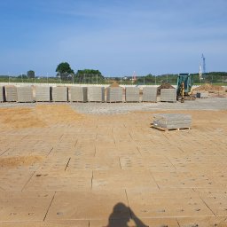 Brukmar Karol Marciński - Przygotowany teren pod kostkę betonową z ułożonymi płytami bazowymi, podsypką piaskową, paletami kostki i mini koparką w tle, w słoneczny dzień.