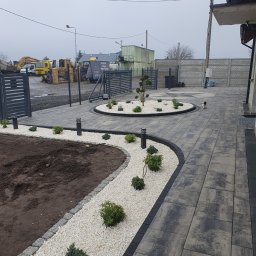 Brukmar Karol Marciński - Ułożona kostka brukowa w szarościach z wykończeniem z białego kamienia i nasadzeniami. W tle brama wjazdowa i sprzęt budowlany. Całość w pochmurny dzień.