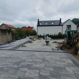 Brukmar Karol Marciński - Układanie szarej kostki brukowej na posesji domu jednorodzinnego, widoczny minikoparka, częściowo ułożona nawierzchnia, przygotowane materiały i narzędzia.