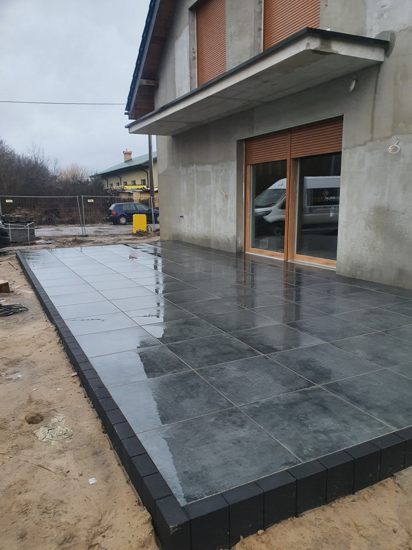 Świeżo ułożona, grafitowa nawierzchnia z kwadratowych płyt, obramowana czarną kostką. W tle budynek w stanie surowym i częściowo widoczne ogrodzenie.