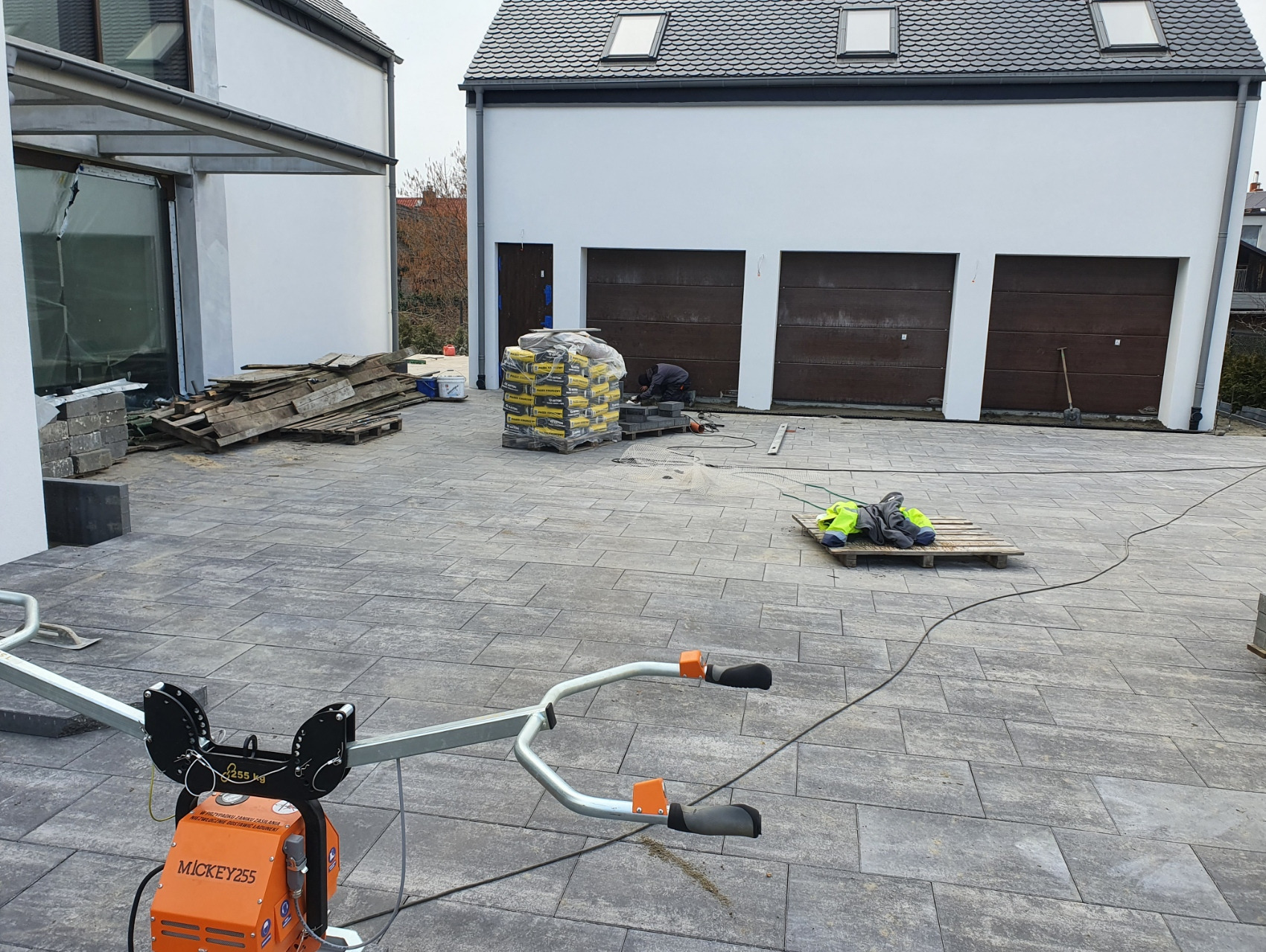 Ukończona nawierzchnia z szarej kostki w Rumi. Widoczna zagęszczarka, paleta z kostką i pracownik w trakcie pracy. Nowoczesna architektura w tle.