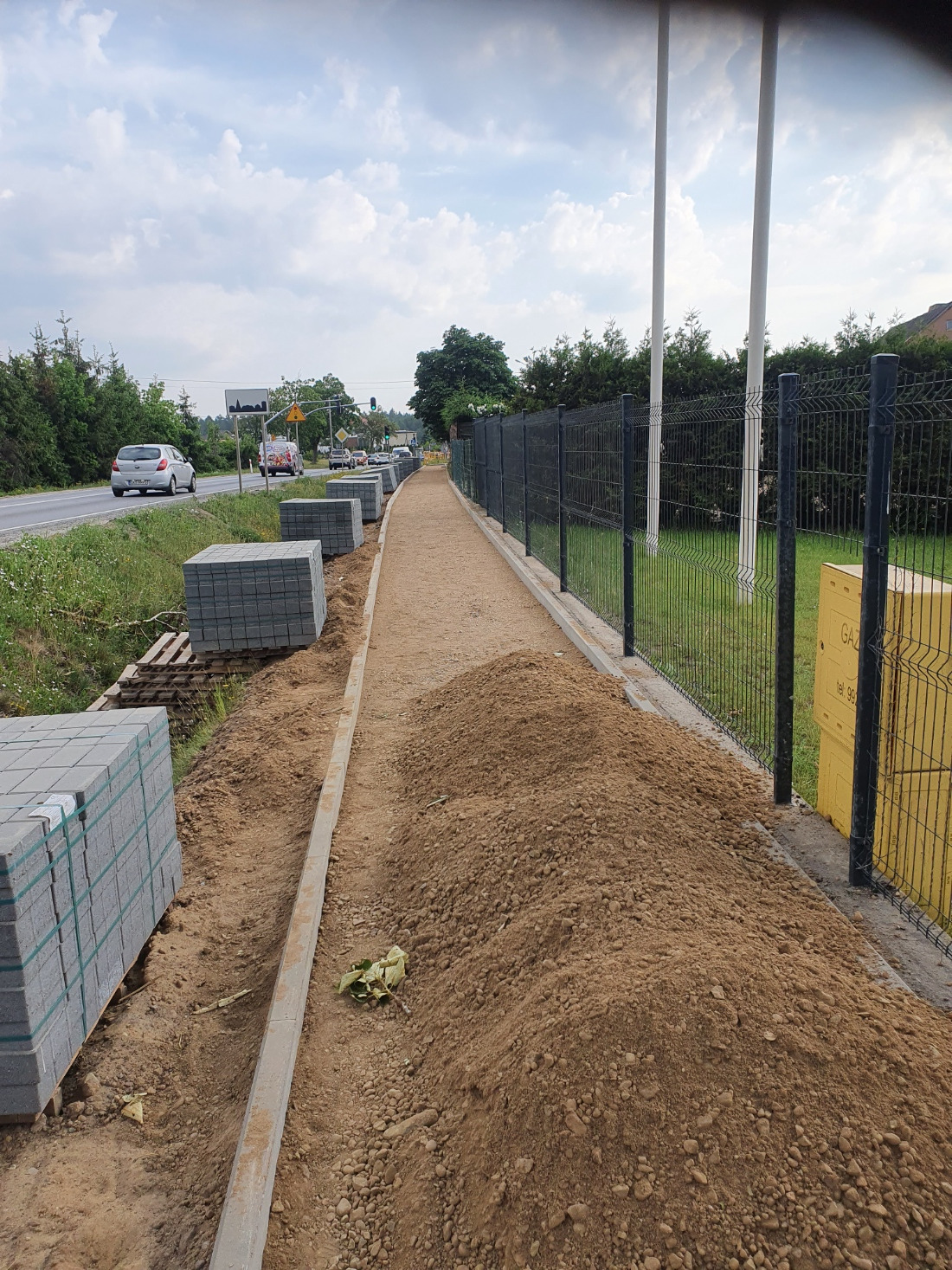 Budowa chodnika: przygotowany teren, ułożone obrzeża, kostka na paletach, montaż ogrodzenia panelowego wzdłuż drogi z widokiem na zieleń i przejeżdżające samochody.