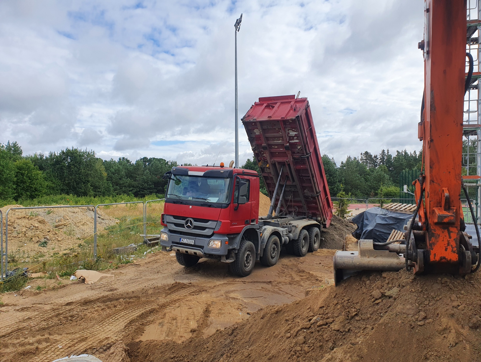 Czerwona wywrotka Mercedes z podniesioną skrzynią ładunkową na placu budowy, obok widoczny pomarańczowy wysięgnik koparki i tymczasowe ogrodzenie.
