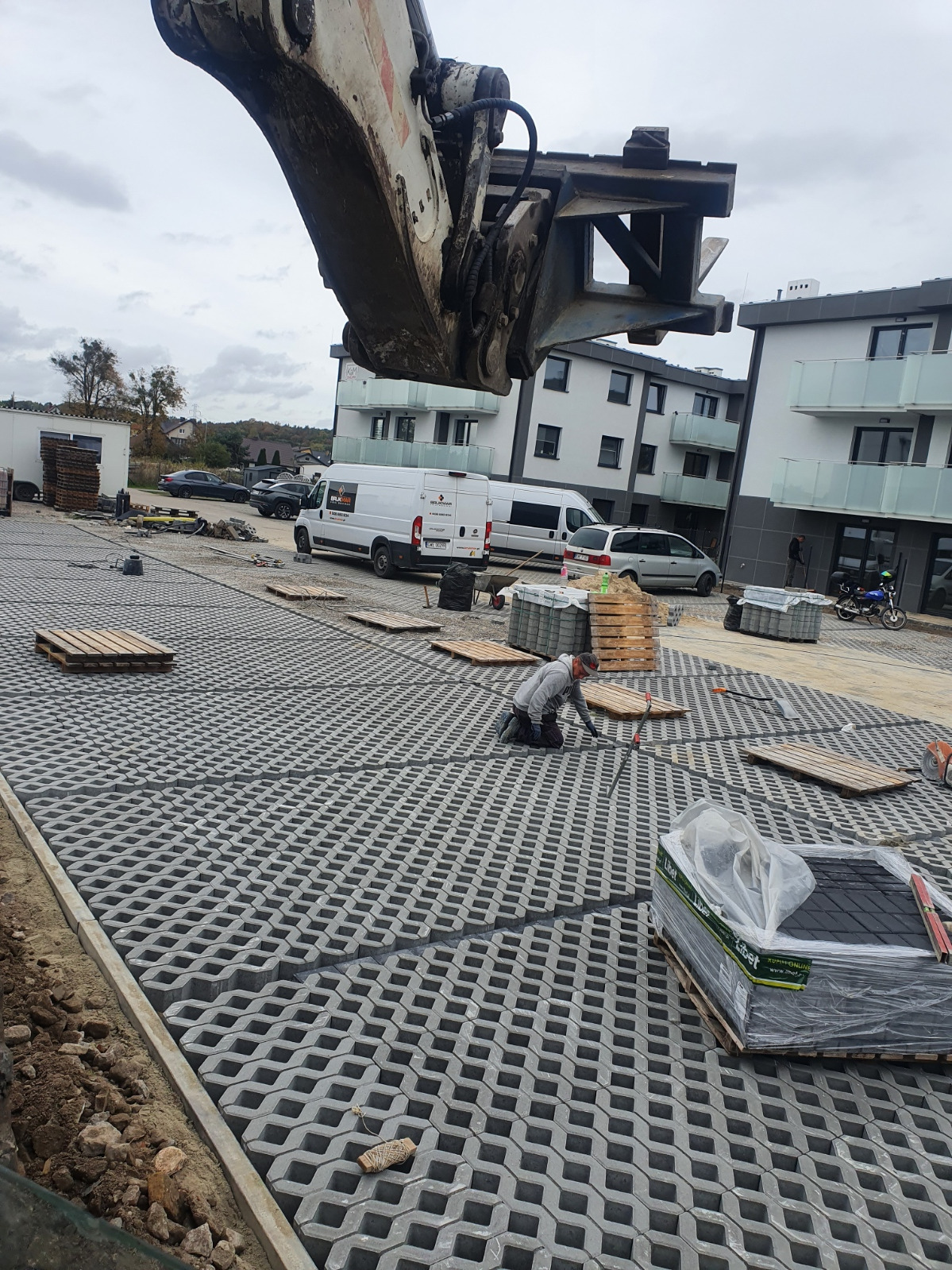 Montaż ażurowej nawierzchni parkingowej z użyciem ciężkiego sprzętu, widoczny pracownik w trakcie pracy, palety z kostką, budynki w tle.