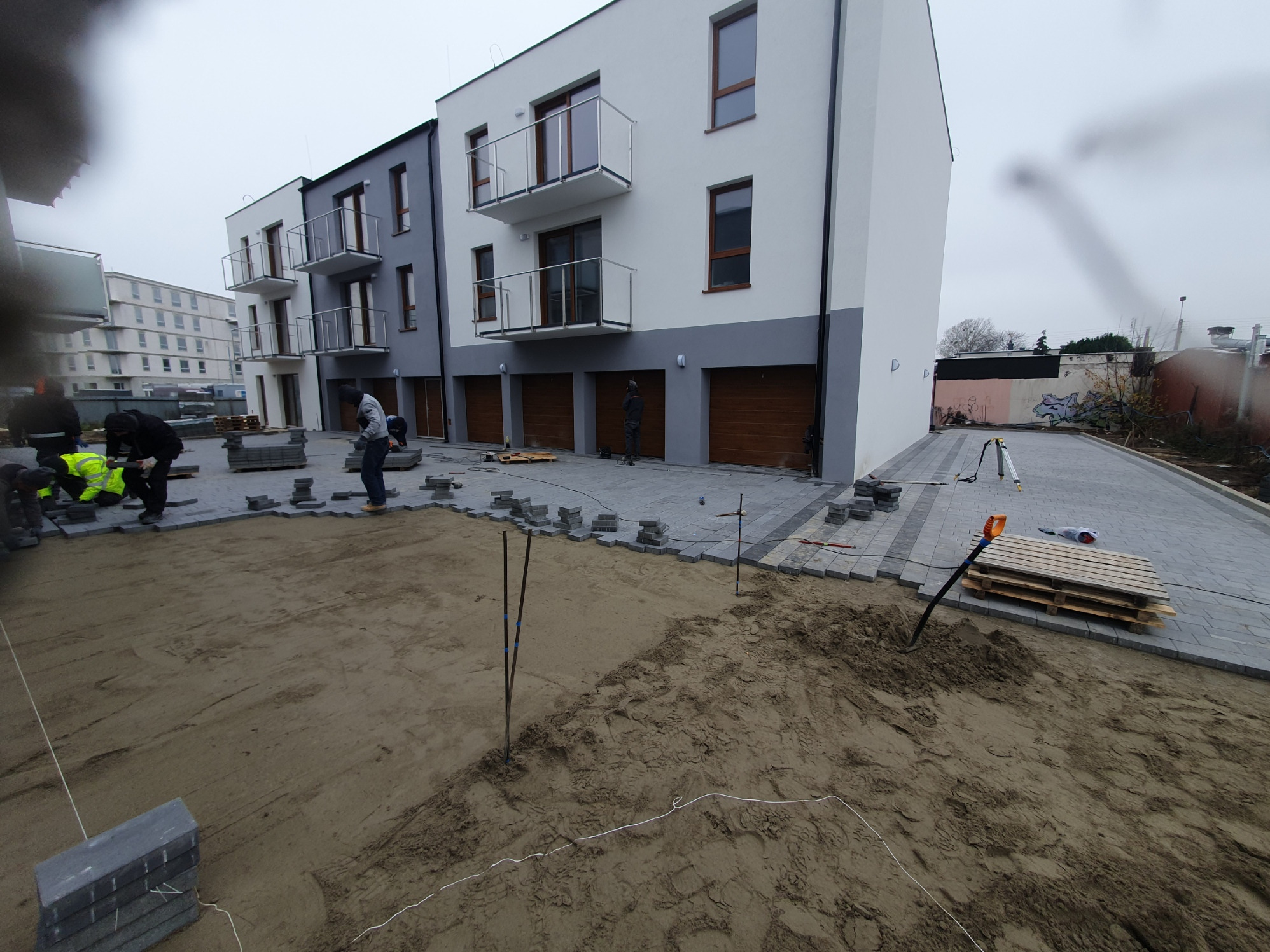 Etap układania szarej kostki brukowej przed budynkiem mieszkalnym z garażami; widoczne narzędzia i pracownicy w odblaskowych kamizelkach. Część nawierzchni już ułożona.
