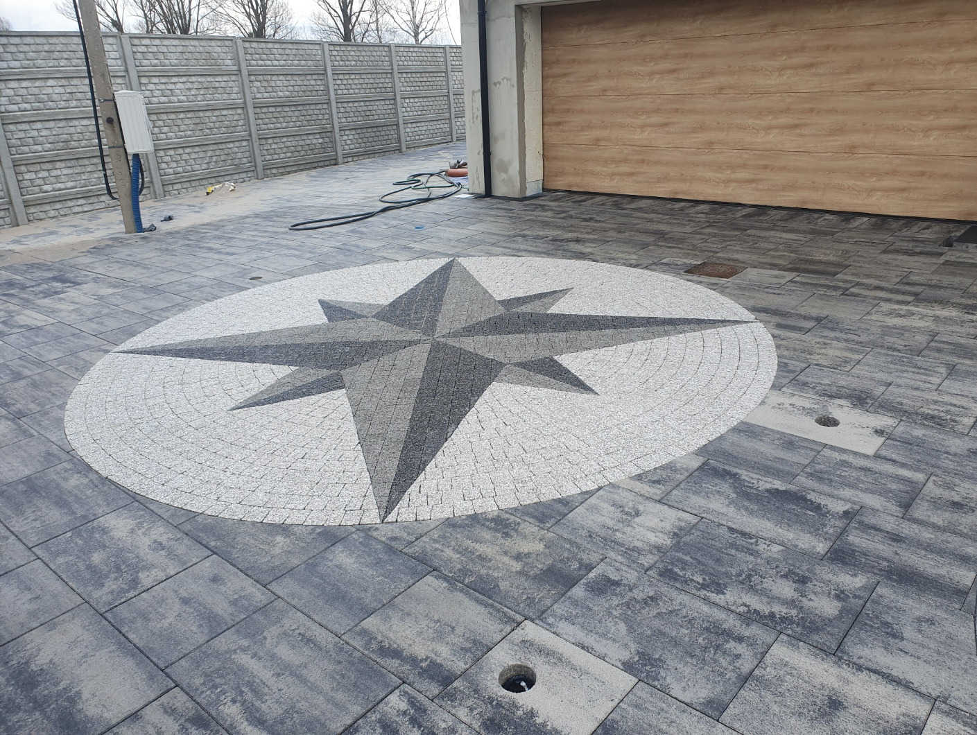 Szara kostka brukowa z dekoracyjnym wzorem kompasu w odcieniach szarości i bieli. W tle widoczny betonowy płot i brama garażowa.