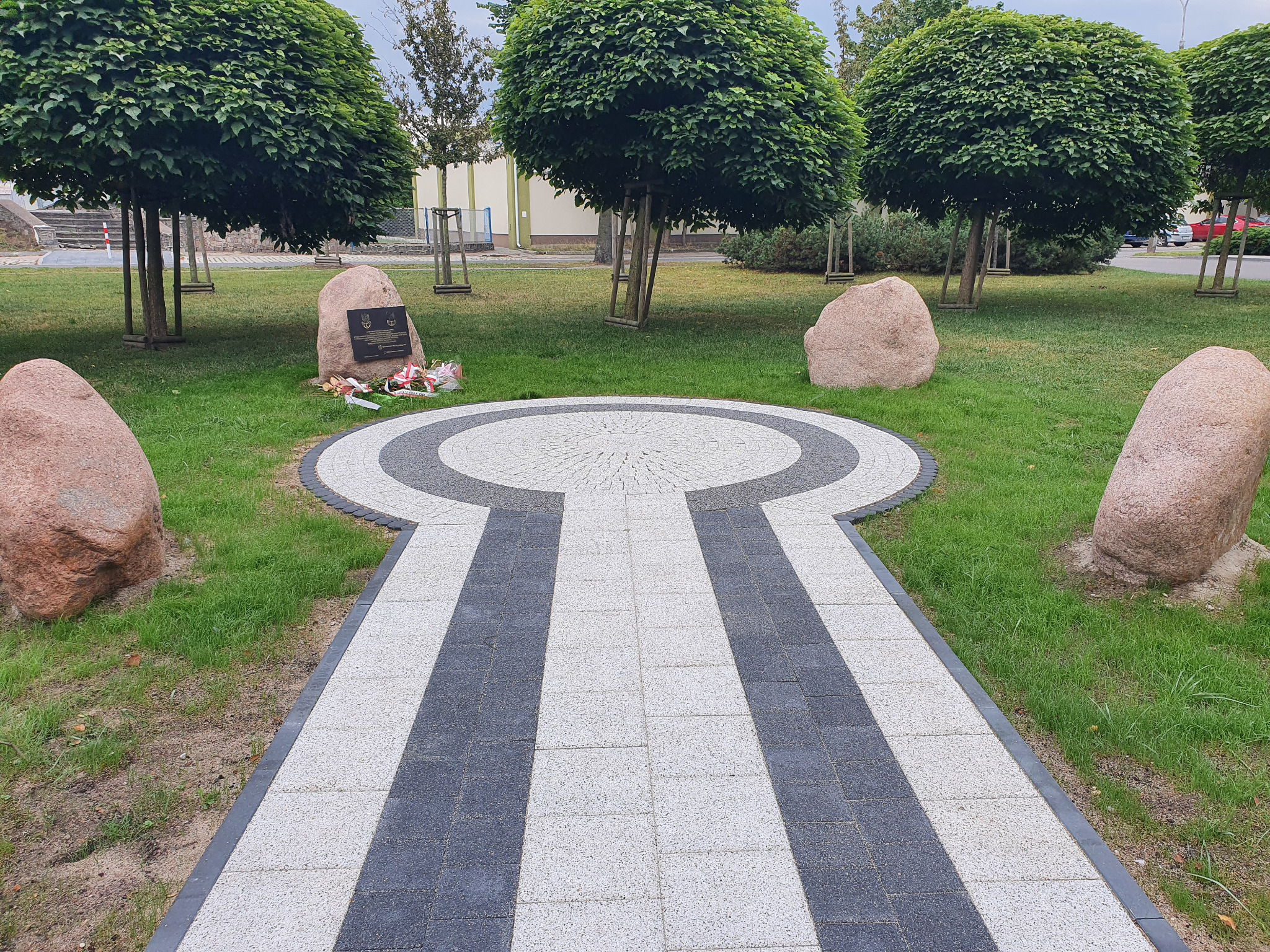 Ozdobna ścieżka z kostki brukowej w parku, prowadząca do pomnika otoczonego kamieniami. Kostka w odcieniach szarości i bieli, drzewa kuliste w tle.