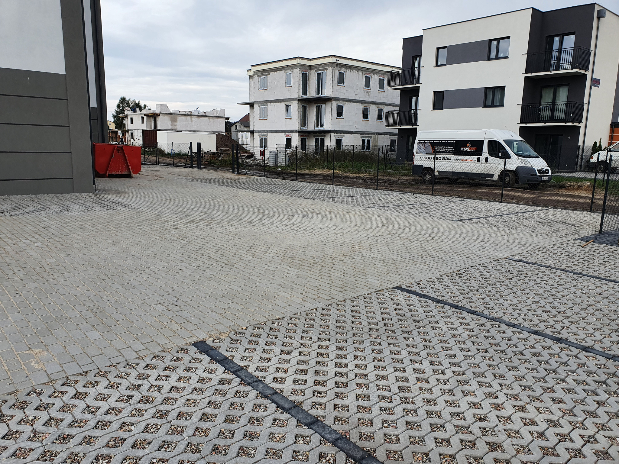 Szara kostka ażurowa i betonowa na posesji nowego bloku. W tle kontener i budynek w trakcie budowy oraz biała furgonetka z logo firmy brukarskiej. Pochmurny dzień.