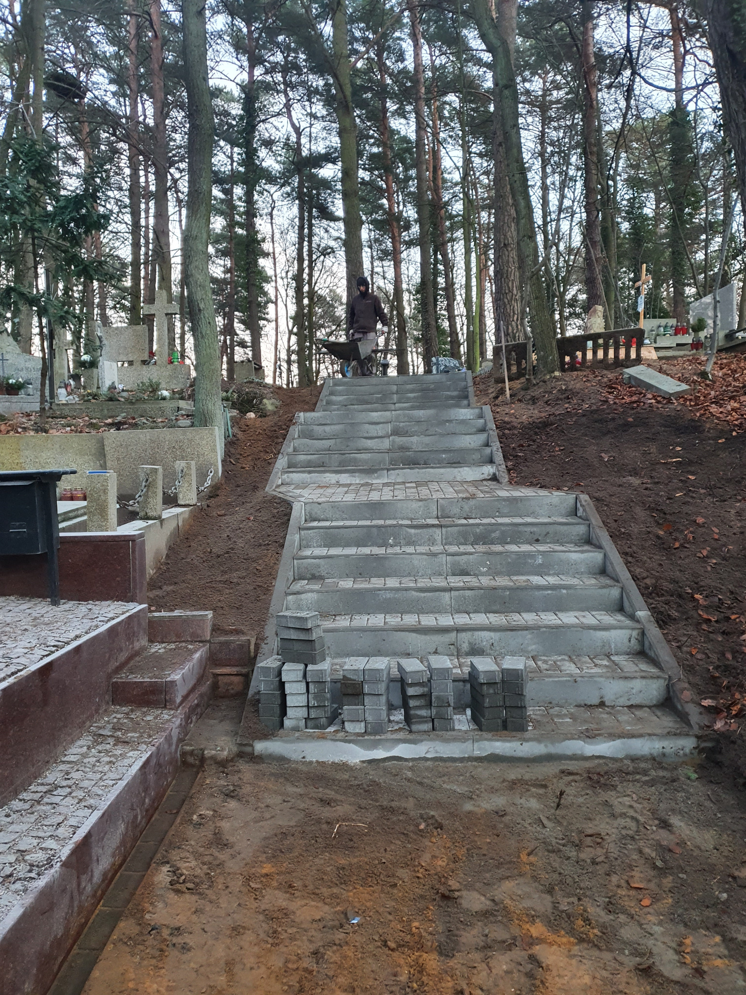 Schody z kostki brukowej w trakcie budowy na cmentarzu, z ułożonymi stopniami i stosem kostki obok. W tle drzewa i nagrobki.