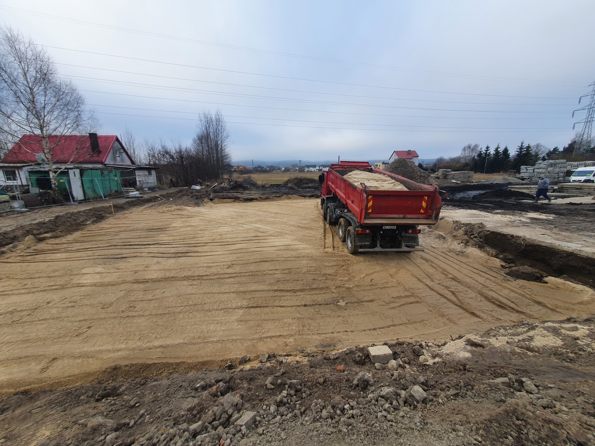 Ciężarówka z piaskiem na wyrównanym terenie budowy, widoczne ślady opon, w tle domy i słupy wysokiego napięcia. Teren przygotowany pod dalsze prace.