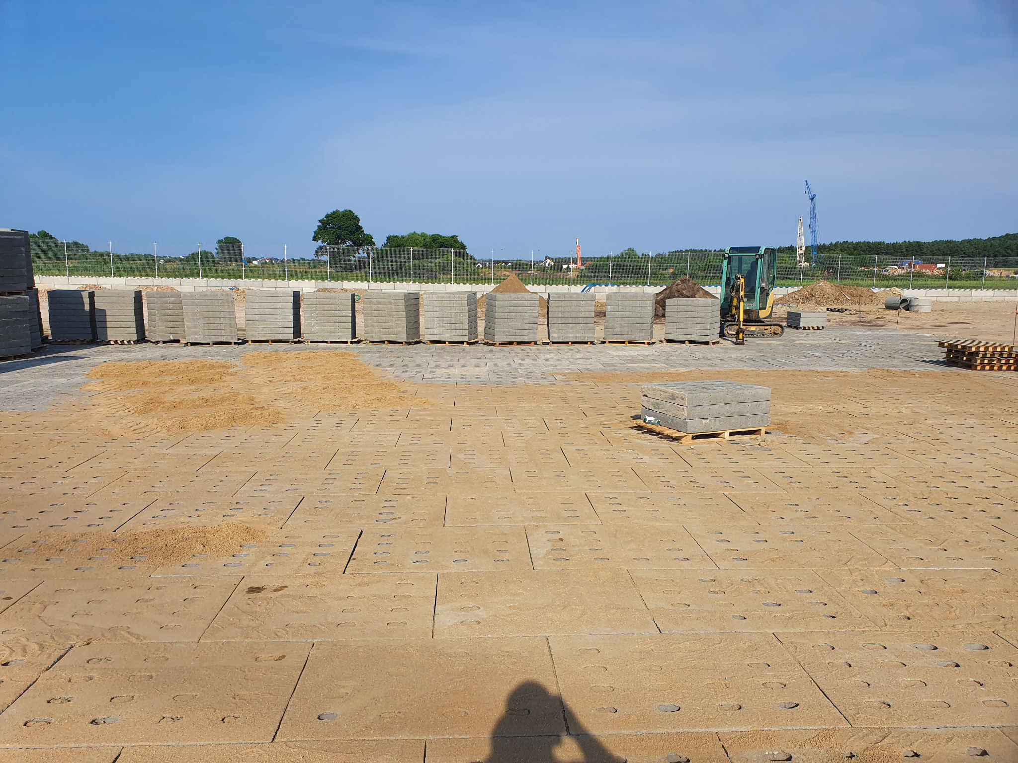 Przygotowany teren pod kostkę betonową z ułożonymi płytami bazowymi, podsypką piaskową, paletami kostki i mini koparką w tle, w słoneczny dzień.