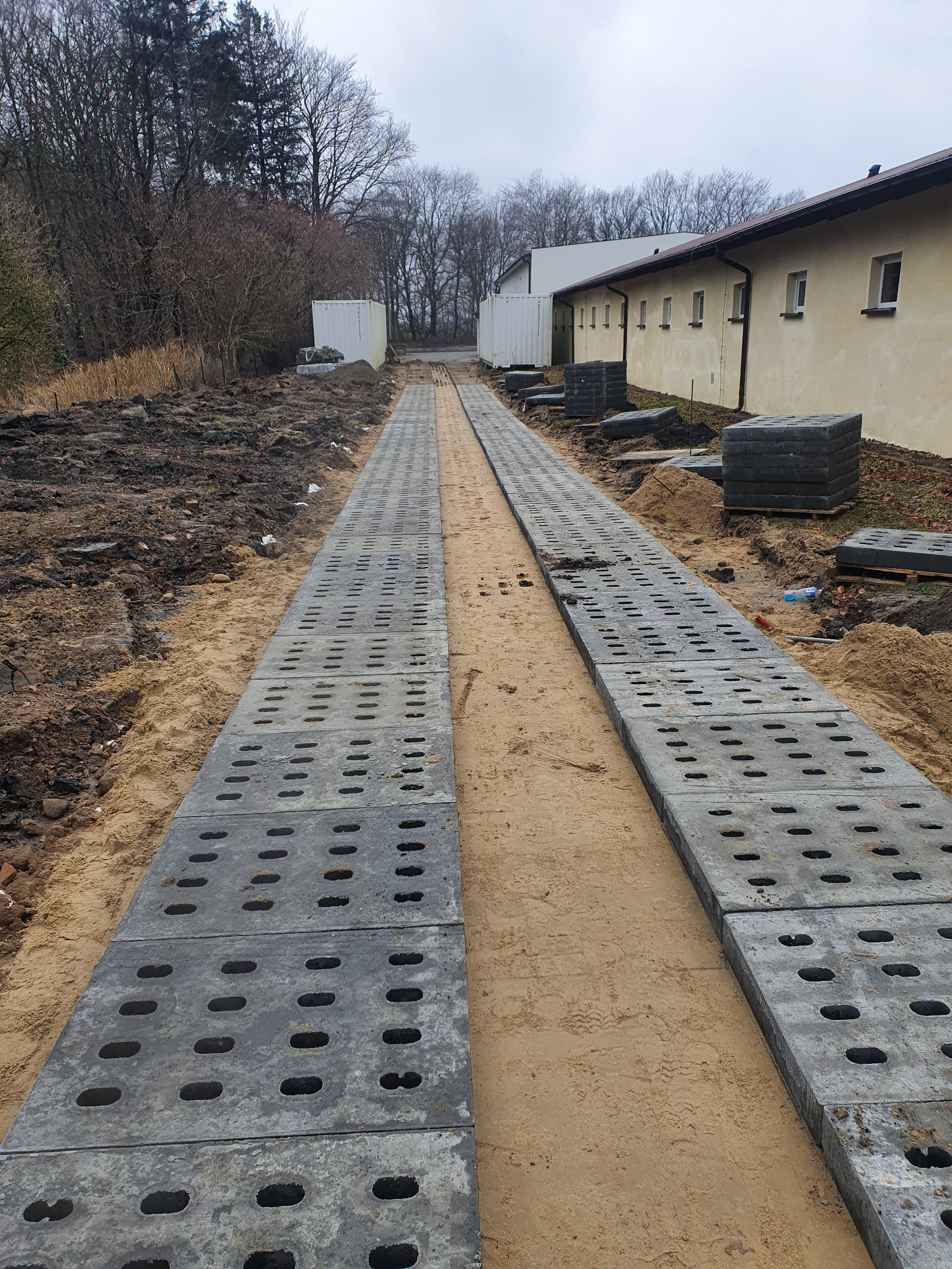 Dwie ścieżki z ażurowych płyt betonowych na piasku, prowadzące wzdłuż budynku. W tle drzewa i kontenery. Widok z perspektywy.