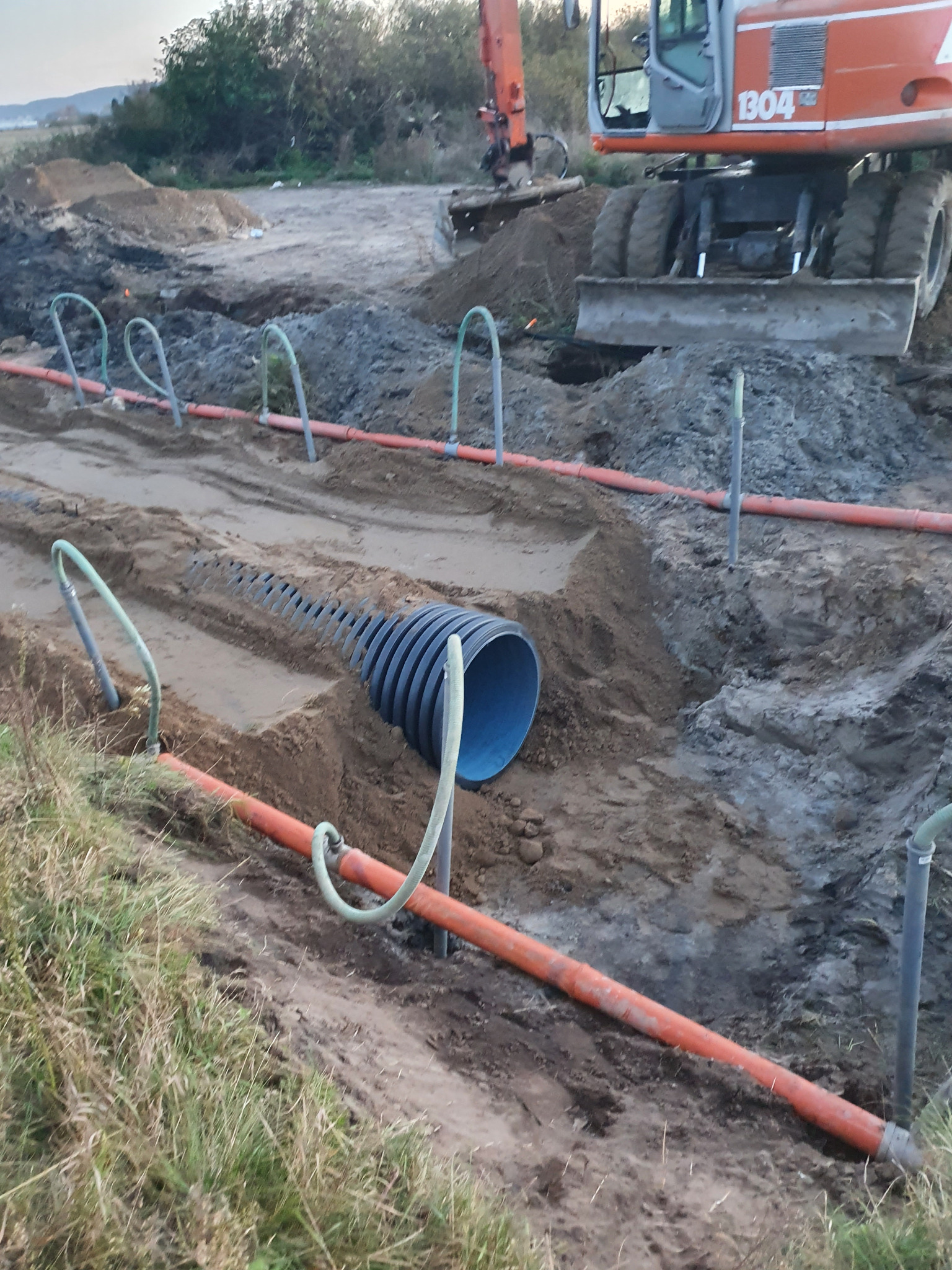 Wykop z ułożonymi rurami drenażowymi i pomarańczowymi rurami, z koparką kołową w tle, na terenie budowy. Widoczne elementy systemu drenażowego i przygotowanie terenu.