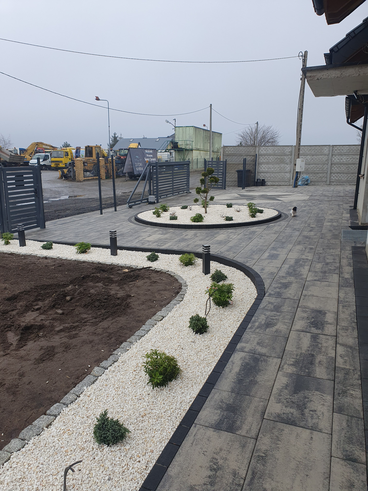 Ułożona kostka brukowa w szarościach z wykończeniem z białego kamienia i nasadzeniami. W tle brama wjazdowa i sprzęt budowlany. Całość w pochmurny dzień.