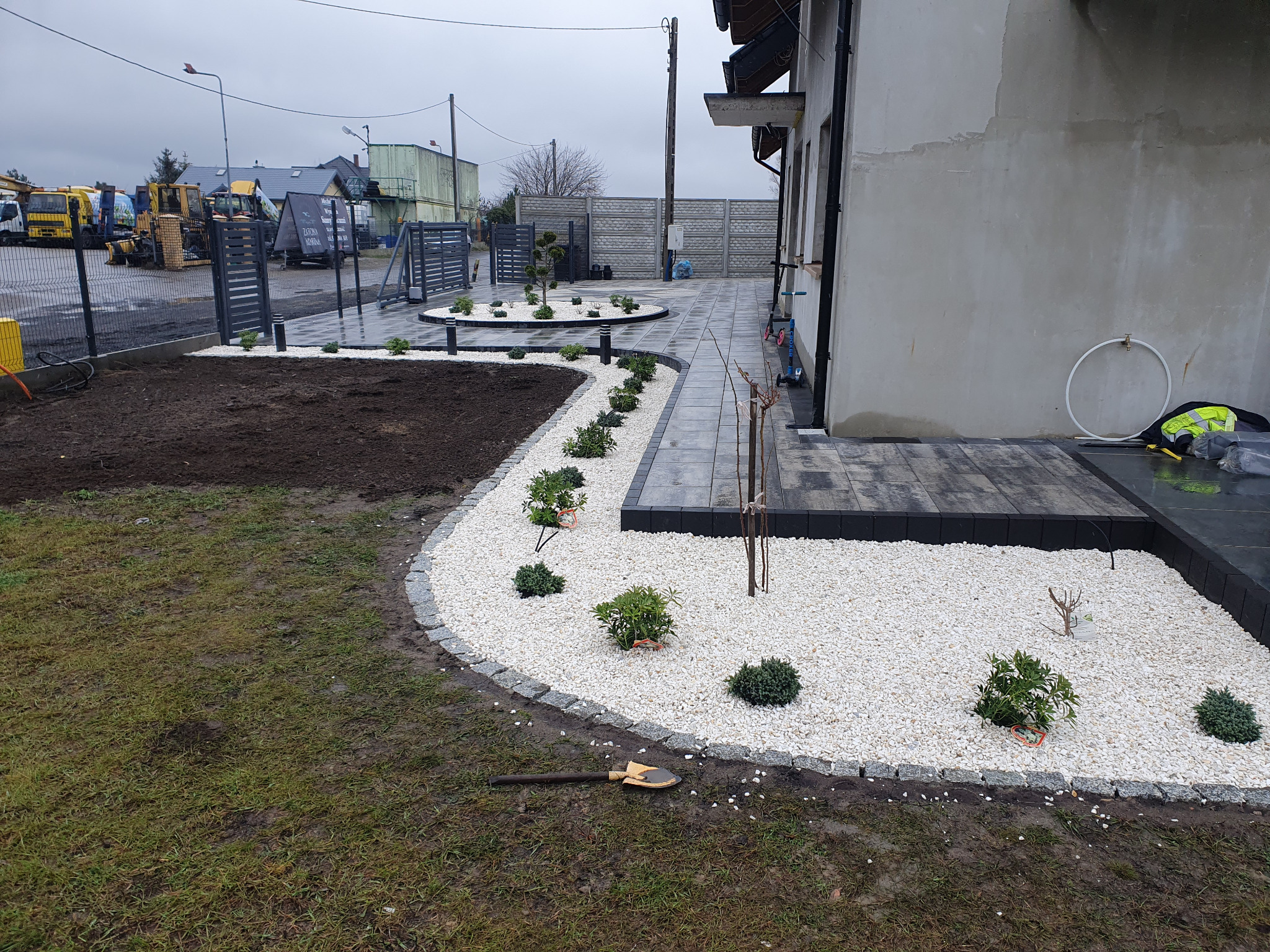 Aranżacja terenu z kostki i białego kamienia wokół domu. Widoczne nasadzenia roślin, oświetlenie i fragment ogrodzenia. W tle sprzęt budowlany.
